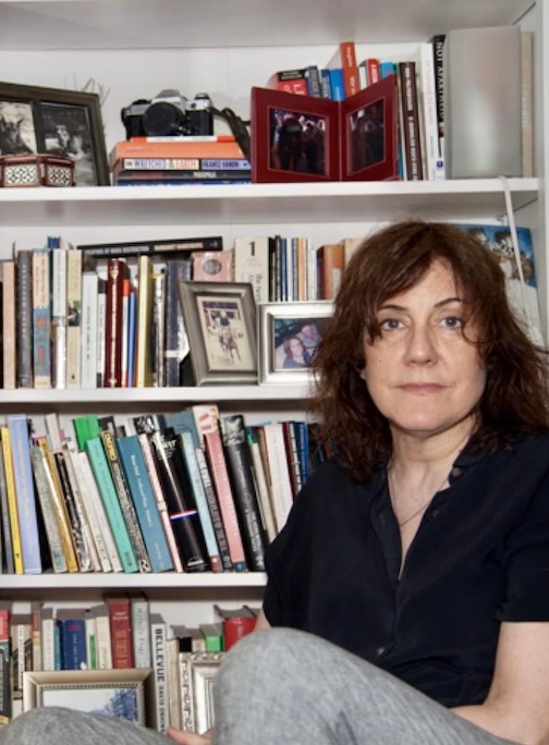 A woman with wavy brown hair and a black shirt sits in front of a bookshelf filled with books, framed photos, and a camera. She looks calmly at the camera.