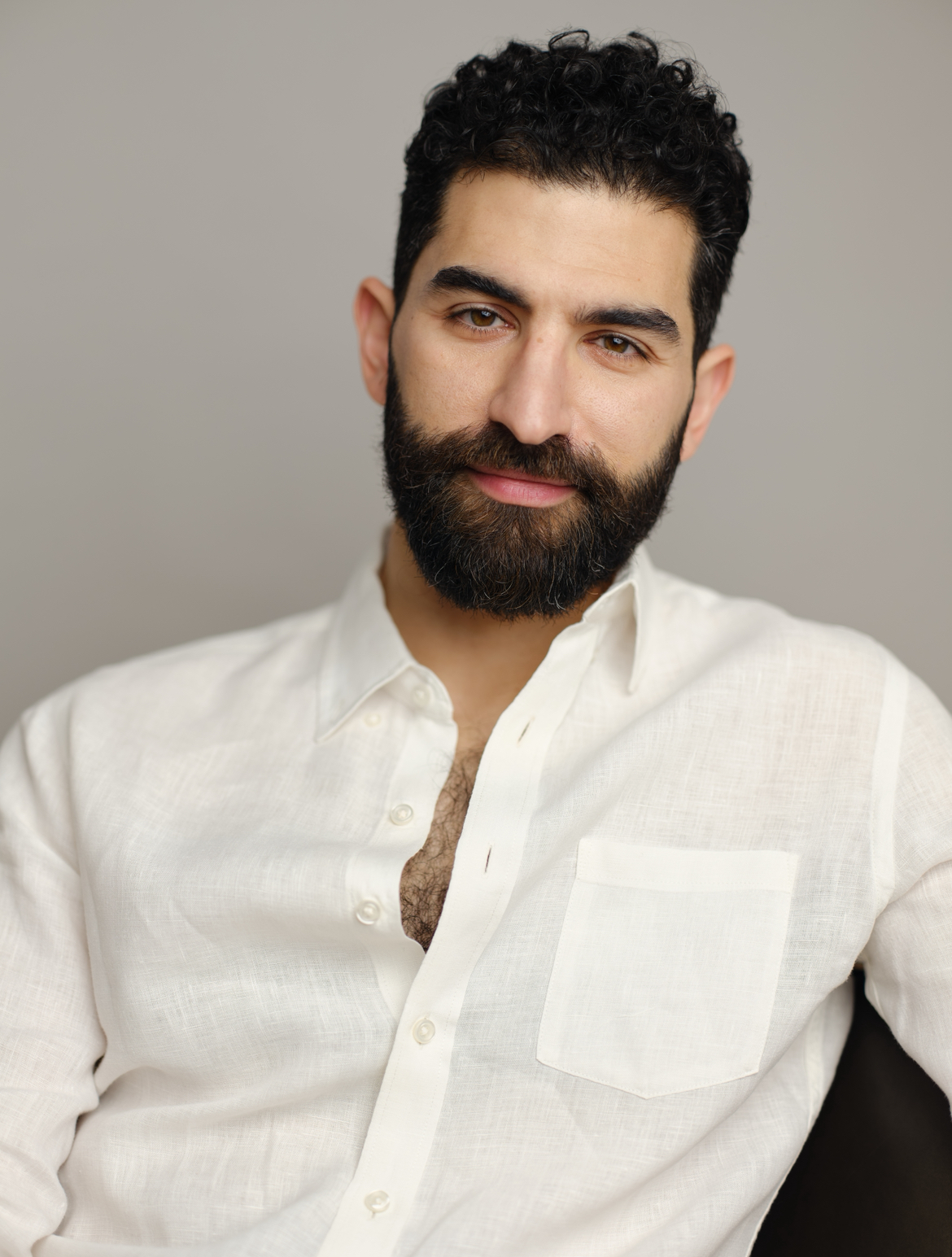 A man with dark curly hair and a full beard wears a white button-up shirt and looks calmly at the camera against a neutral background.
