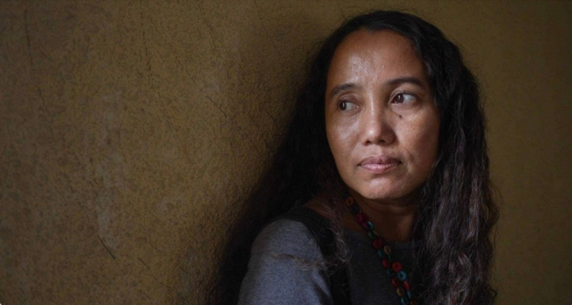 A woman with long, wavy hair wearing a gray top and a beaded necklace gazes thoughtfully to her right while leaning against a textured, yellowish wall.