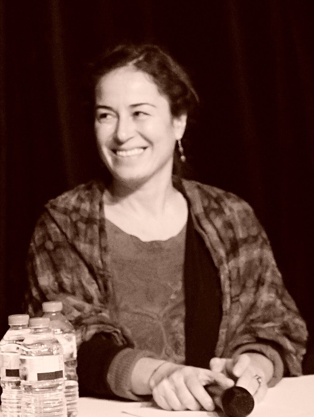 A woman with wavy hair smiles while seated at a table with bottled water in front of her. She holds a microphone and wears a patterned shawl. The image is in sepia tone.