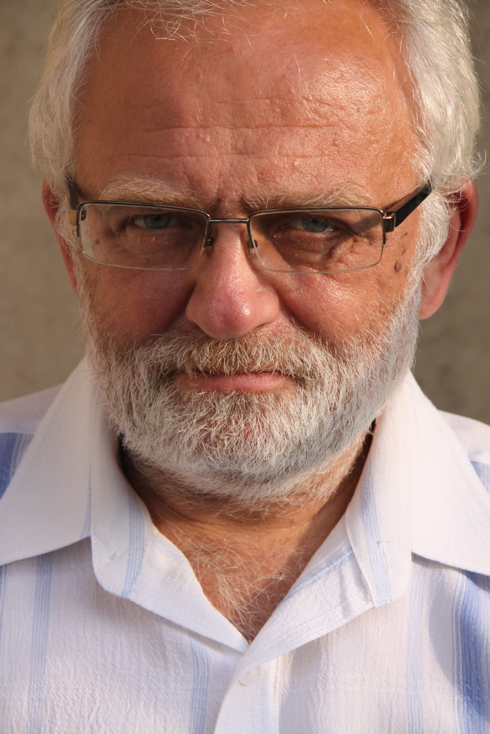 An older man with white hair, a thick white beard, and glasses looks directly at the camera. He is wearing a white, collared shirt with light blue stripes and is standing against a neutral background.