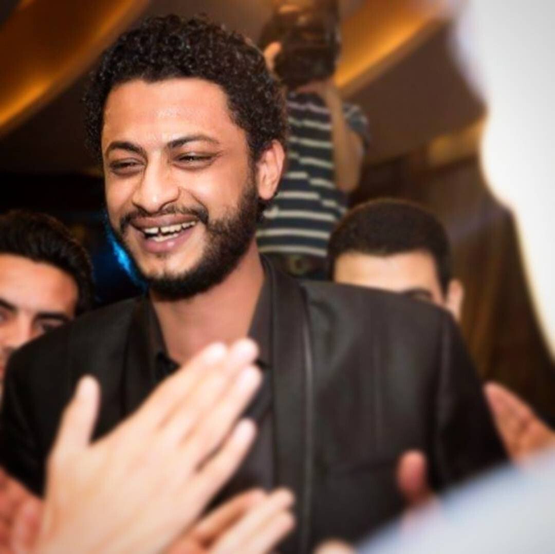A smiling man with curly hair and a beard, wearing a black jacket, is surrounded by people clapping and celebrating. A person in the background is holding a camera and filming the scene.