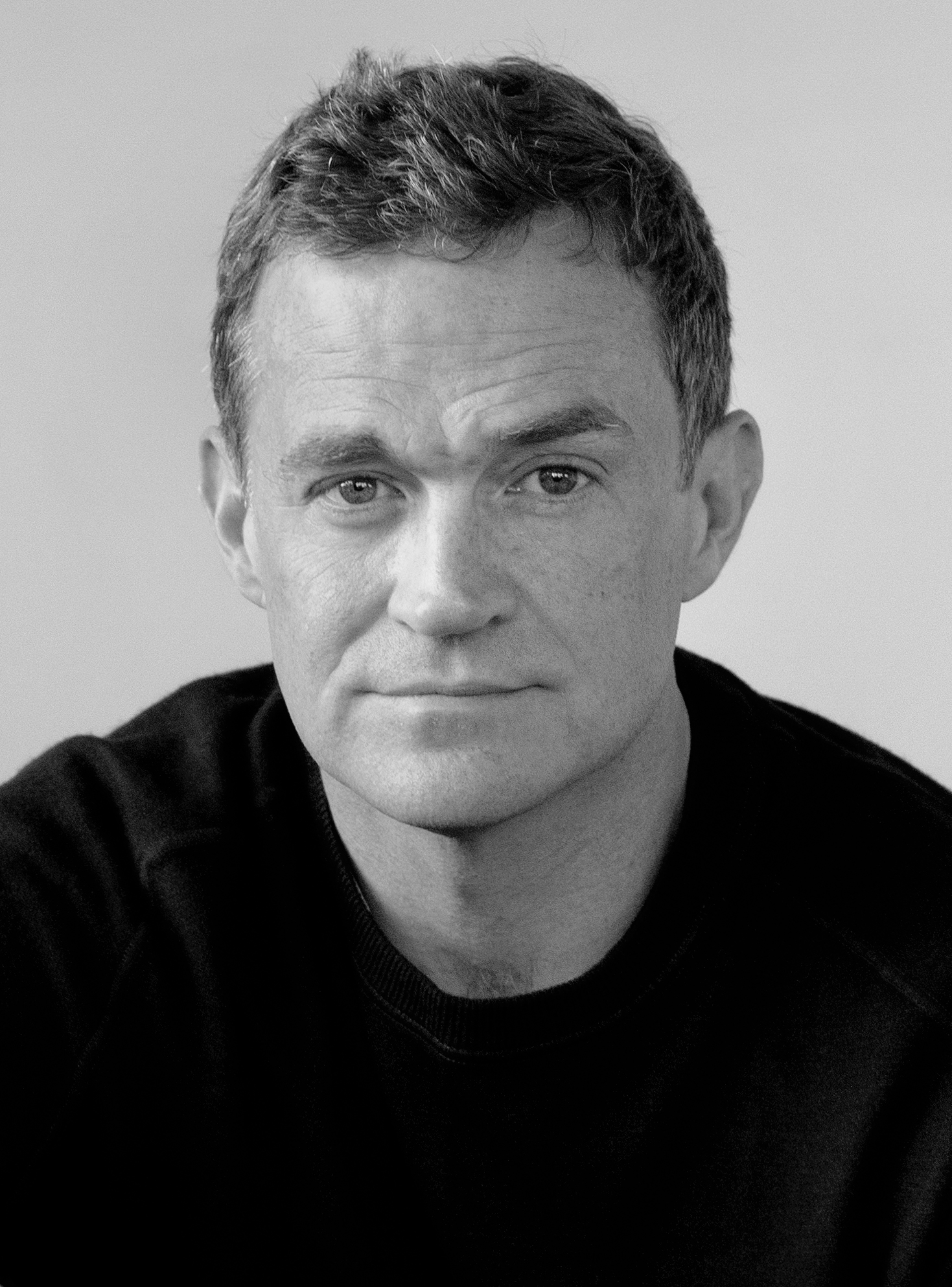 A man with short, wavy hair and a serious expression looks directly at the camera. He is wearing a dark sweater and the background is plain and light-colored. The photo is in black and white.