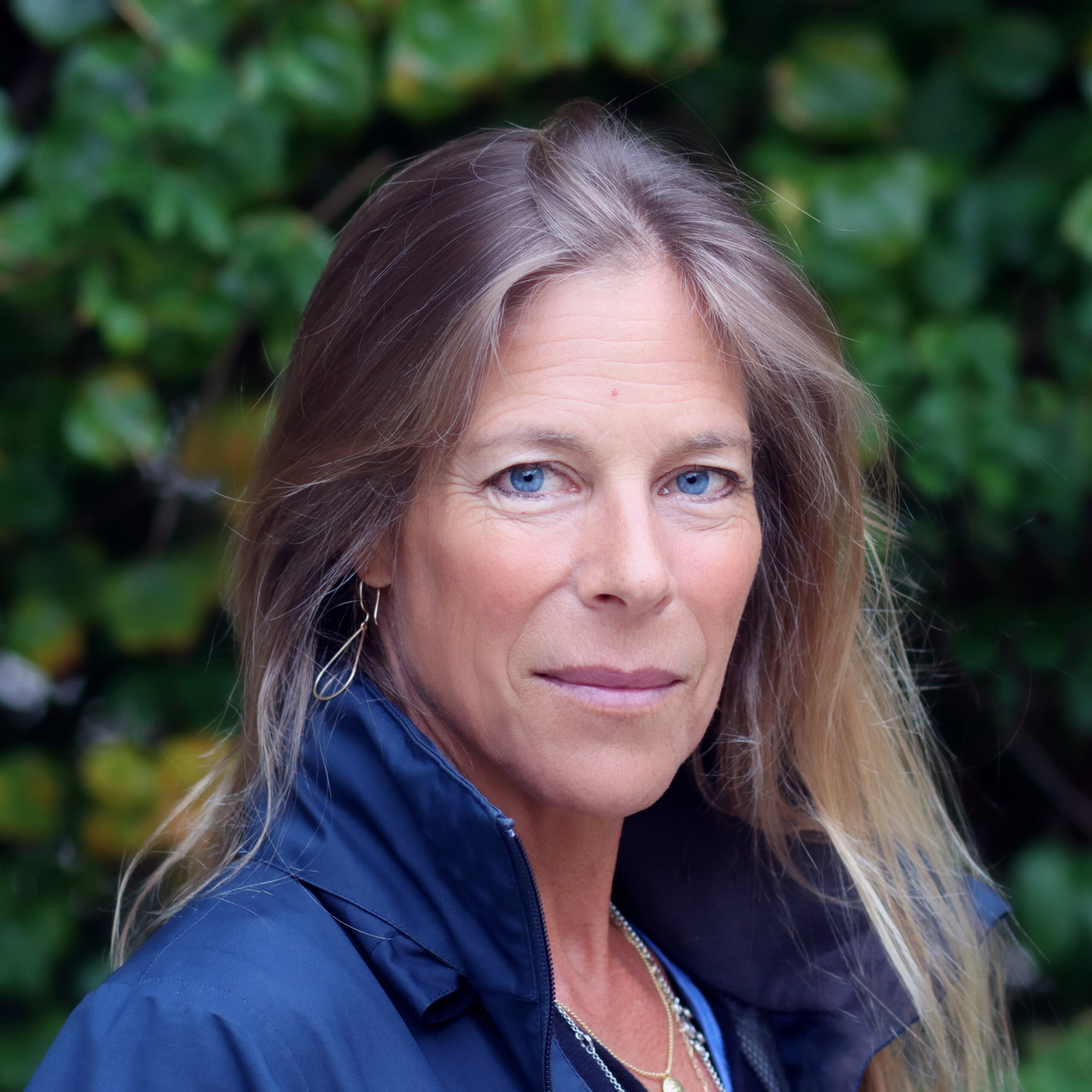 A woman with long blonde hair and blue eyes wearing a dark jacket stands outdoors in front of leafy green foliage, looking directly at the camera with a neutral expression.