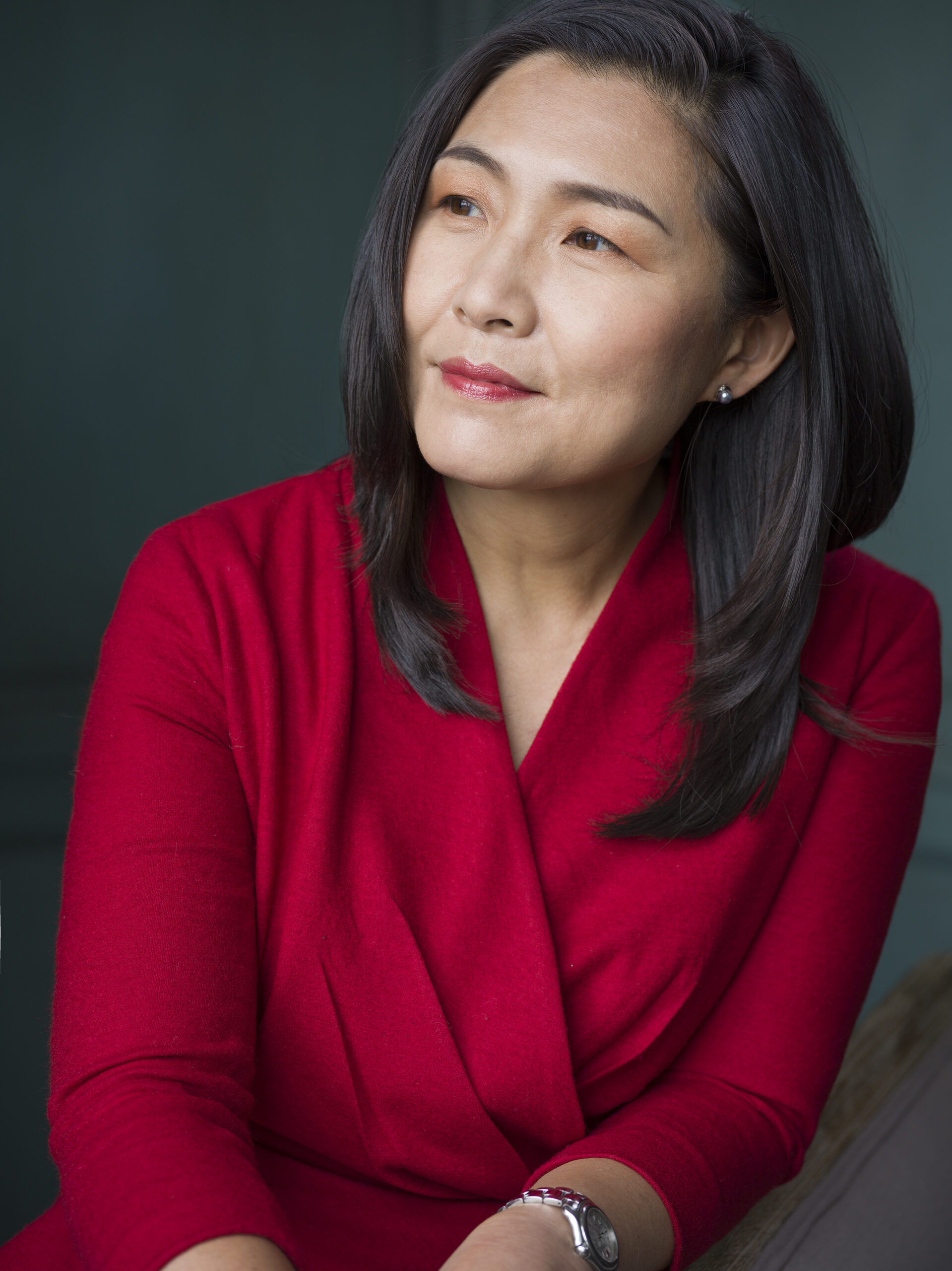 A woman with straight dark hair wearing a red wrap dress looks thoughtfully into the distance against a dark green background. She has a neutral expression and wears a watch on her wrist.