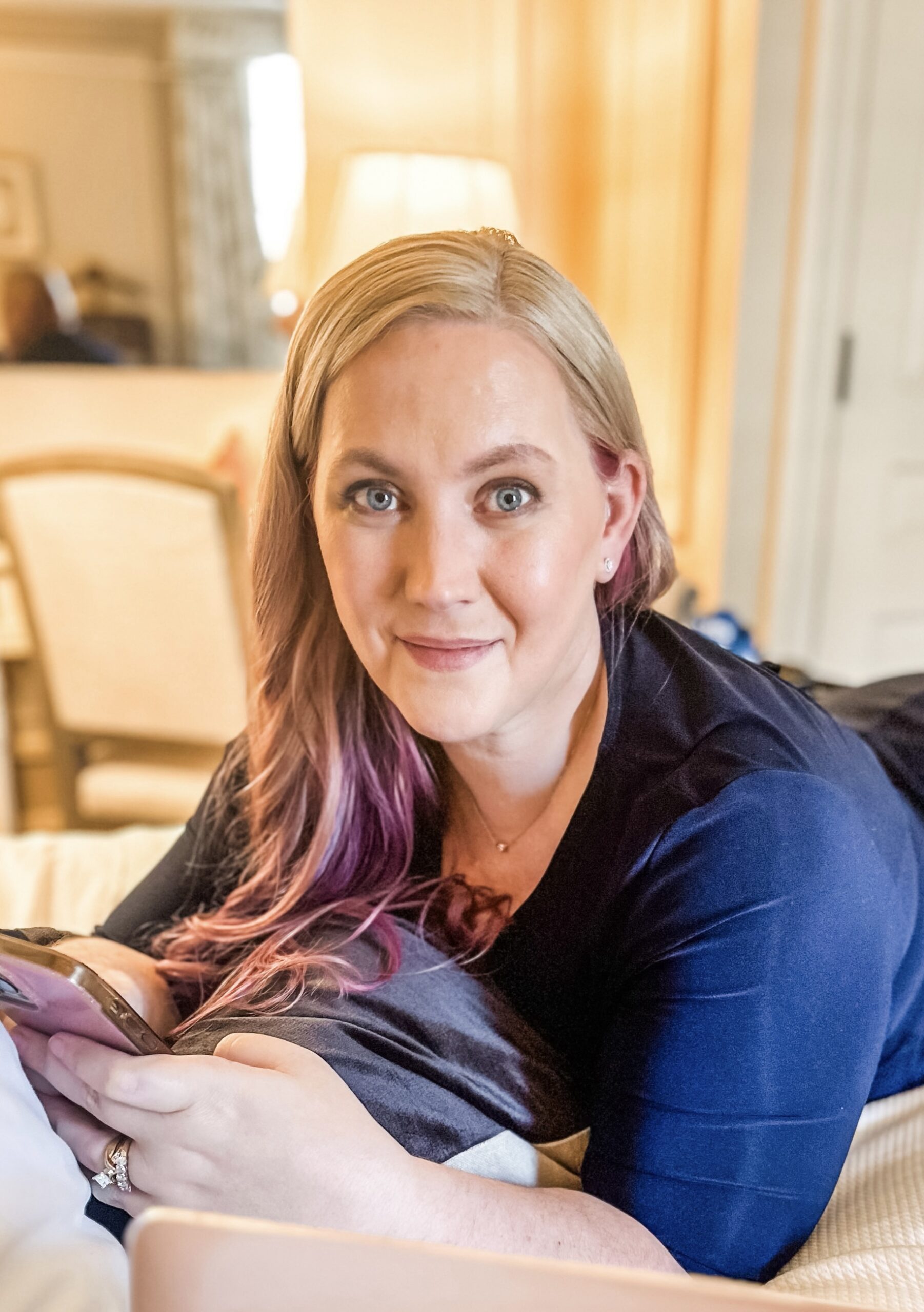 A woman with long, blonde hair with pink tips is lying on a bed, holding a phone and smiling at the camera. She is wearing a navy blue top, and the room is softly lit with warm, natural light.