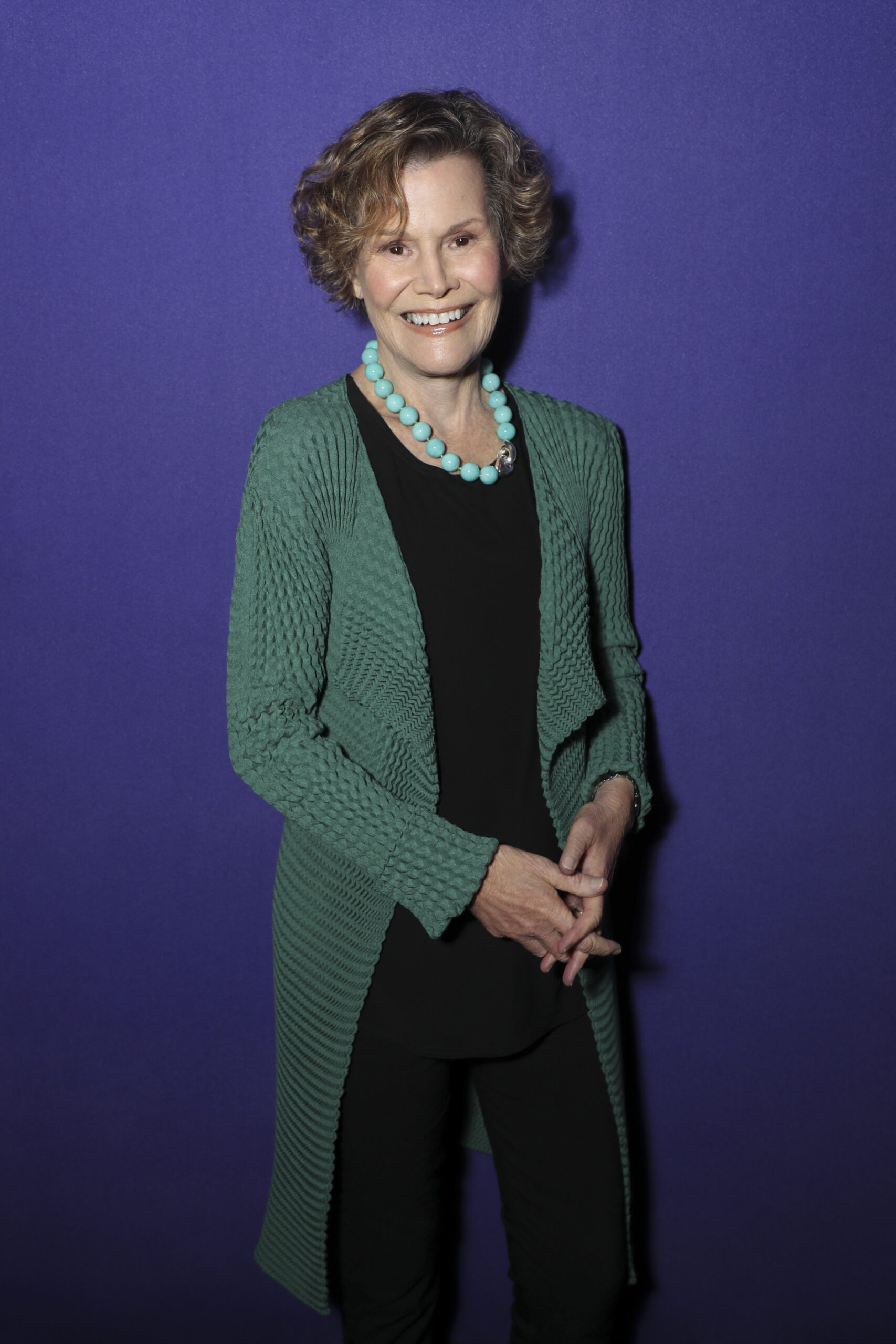 A woman with short curly hair smiles, standing against a solid purple background. She wears a long green cardigan, black top and pants, and a chunky turquoise necklace. Her hands are loosely clasped in front of her.
