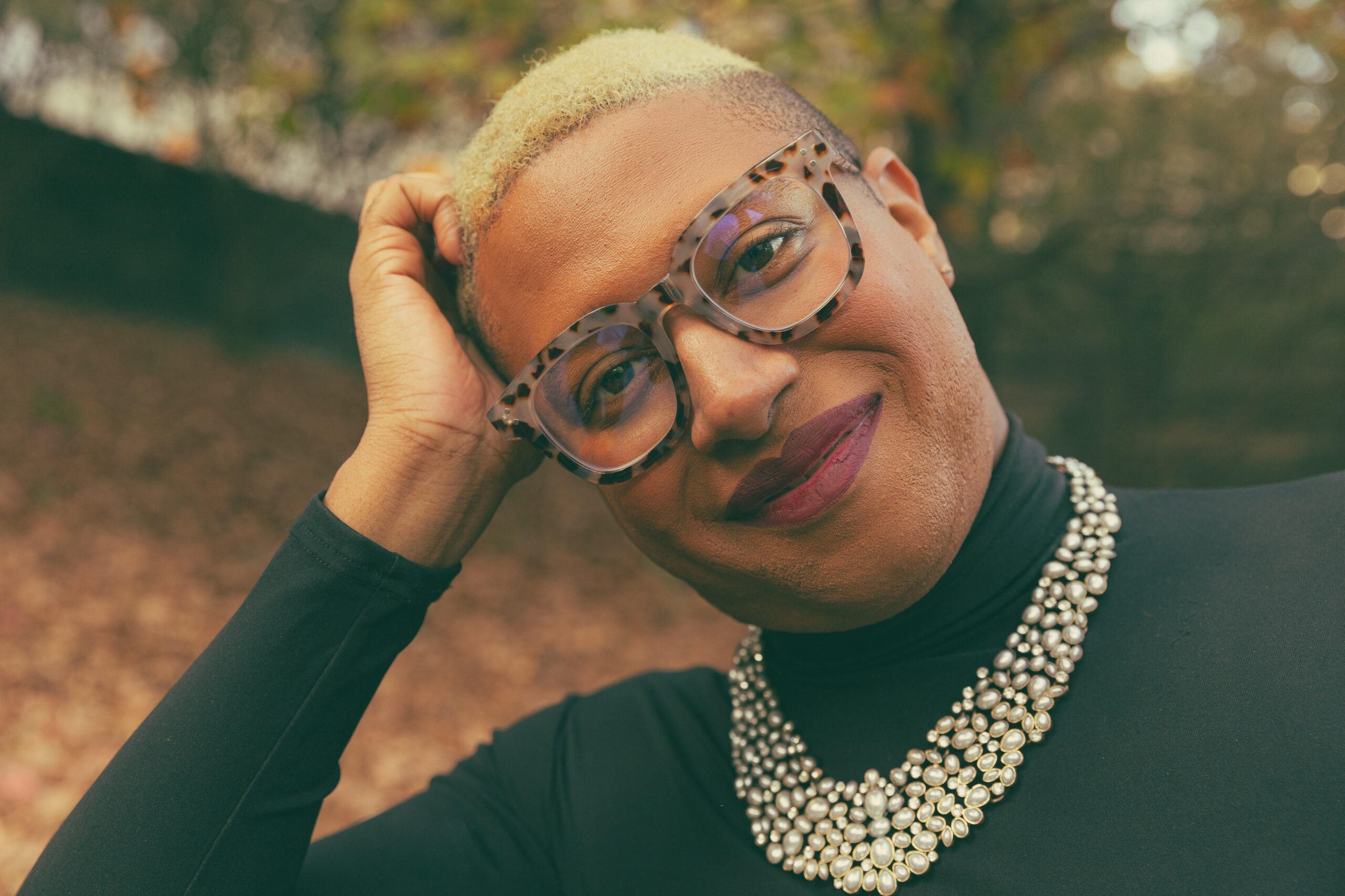 A person with short blond hair, tortoiseshell glasses, and a pearl necklace smiles gently while tilting their head, standing outdoors with blurred autumn foliage in the background.