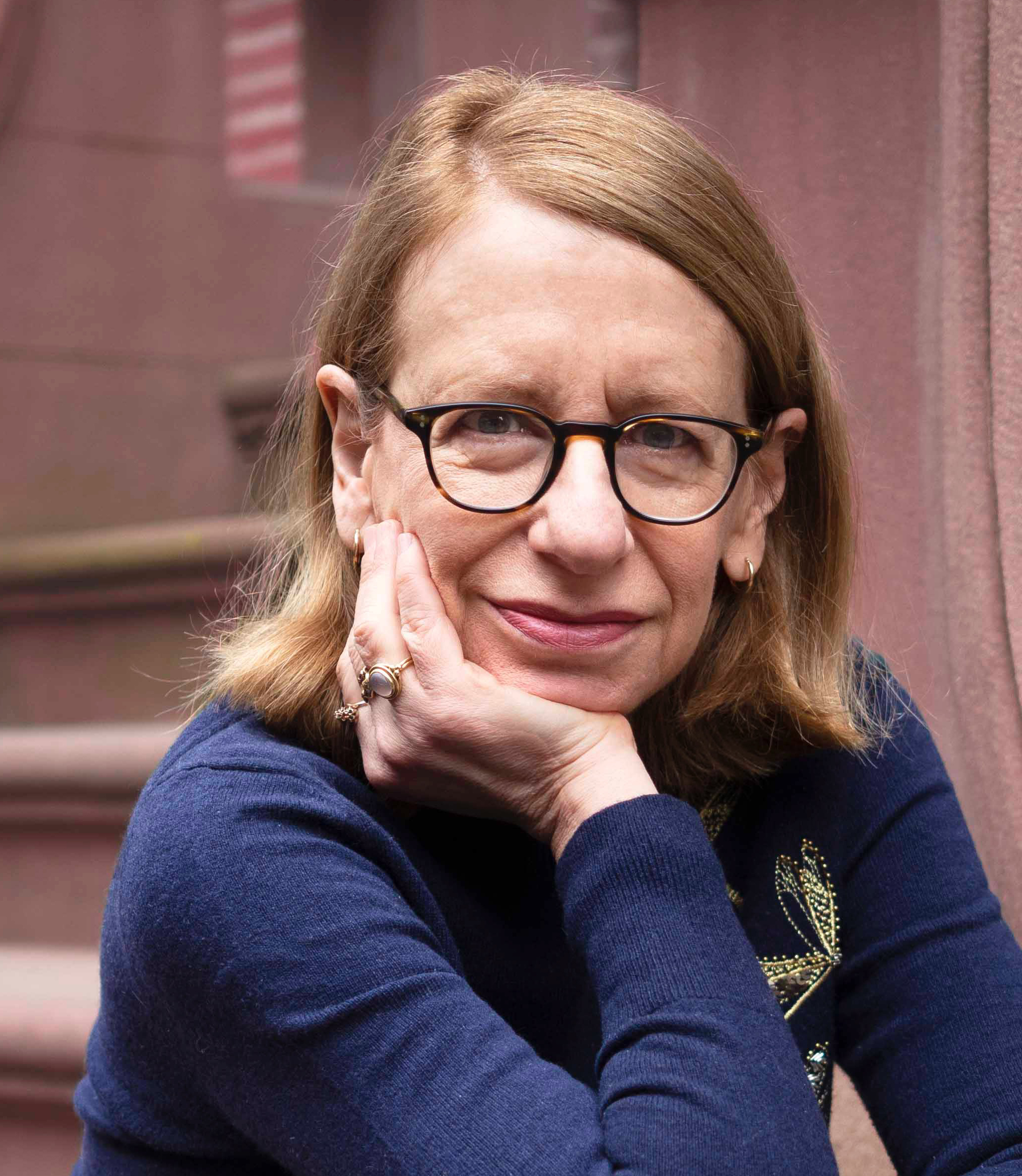 A woman with shoulder-length light brown hair and glasses rests her chin on her hand, smiling slightly. She is wearing a navy blue sweater and sitting in front of brownstone steps.