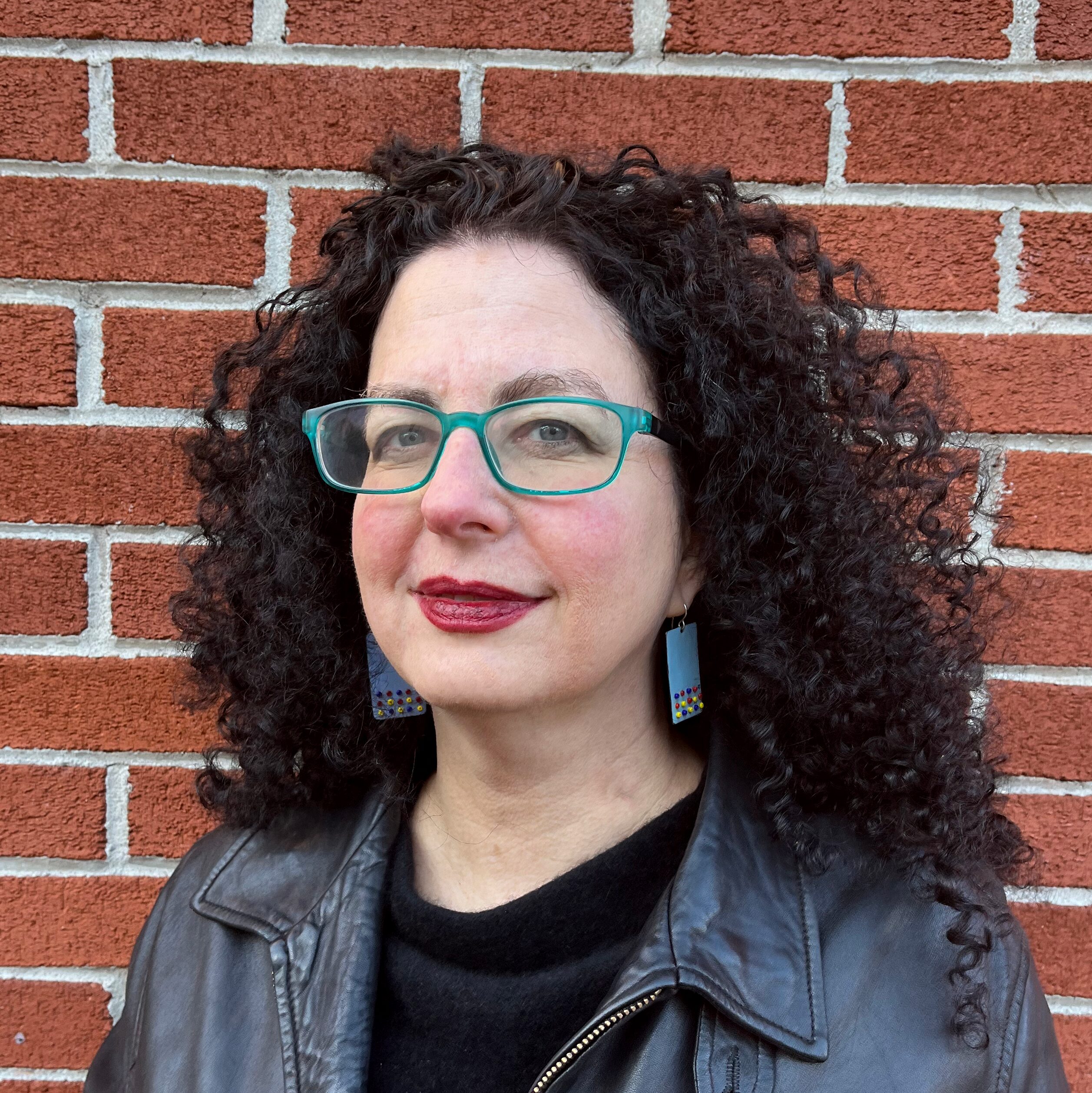 Woman with curly dark hair, teal glasses, and colorful earrings stands in front of a red brick wall, wearing a black leather jacket and dark top. She is looking slightly to the side with a neutral expression.