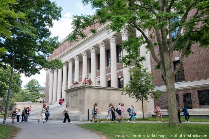 Students walk outside a large university building with tall columns and red brick walls, surrounded by trees on a sunny day. Some people are gathered on steps and a raised platform near the entrance.