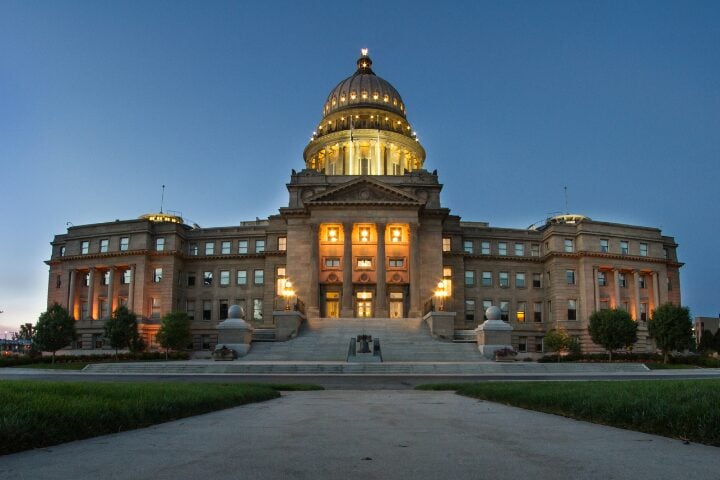 A majestic, illuminated capitol building with a prominent dome stands at dusk. The structure features columns and steps leading up to the entrance, with trees flanking both sides, under a clear, darkening sky.