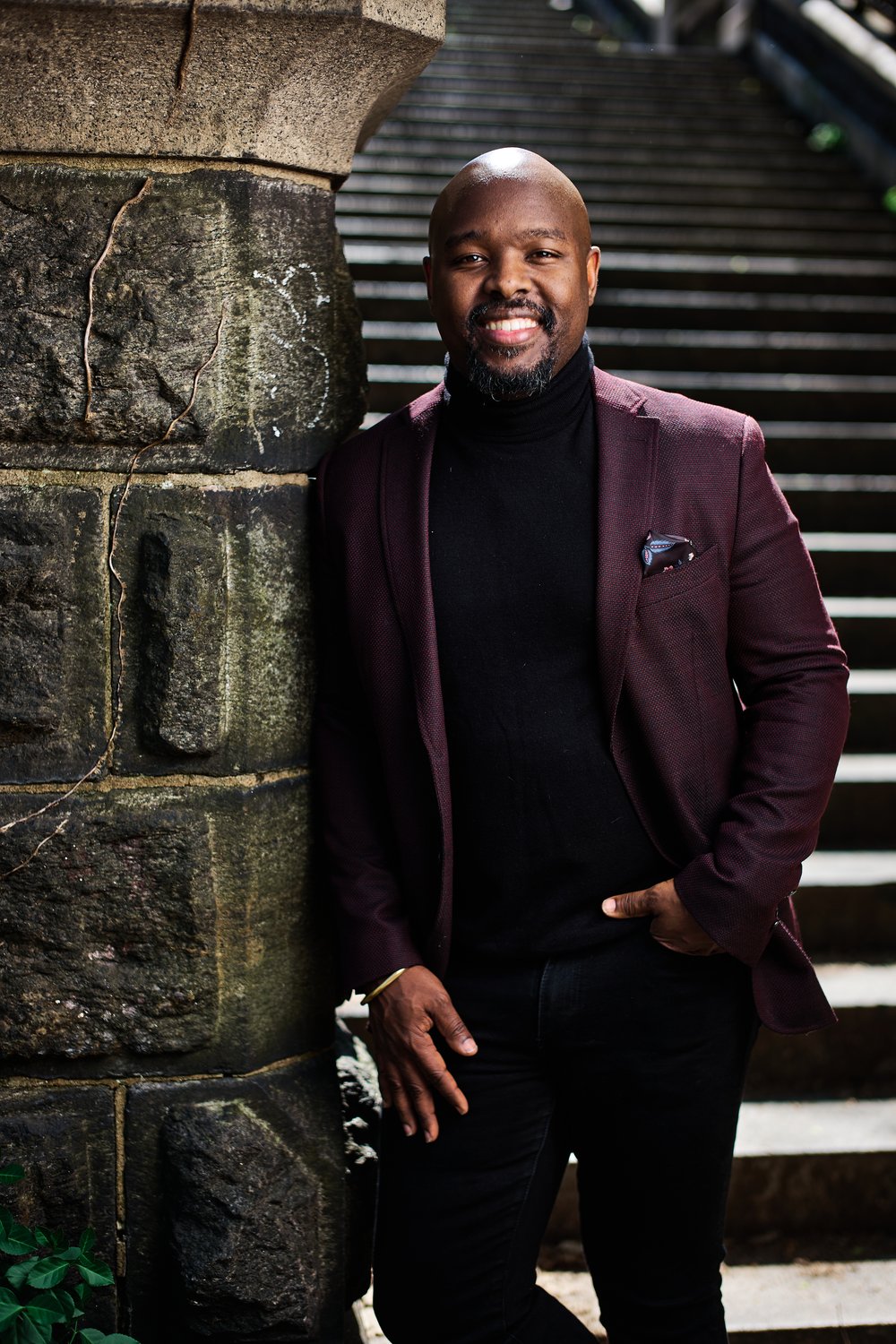 A man in a burgundy blazer and black turtleneck stands against a stone wall, smiling. There are stone steps in the background, leading upward.