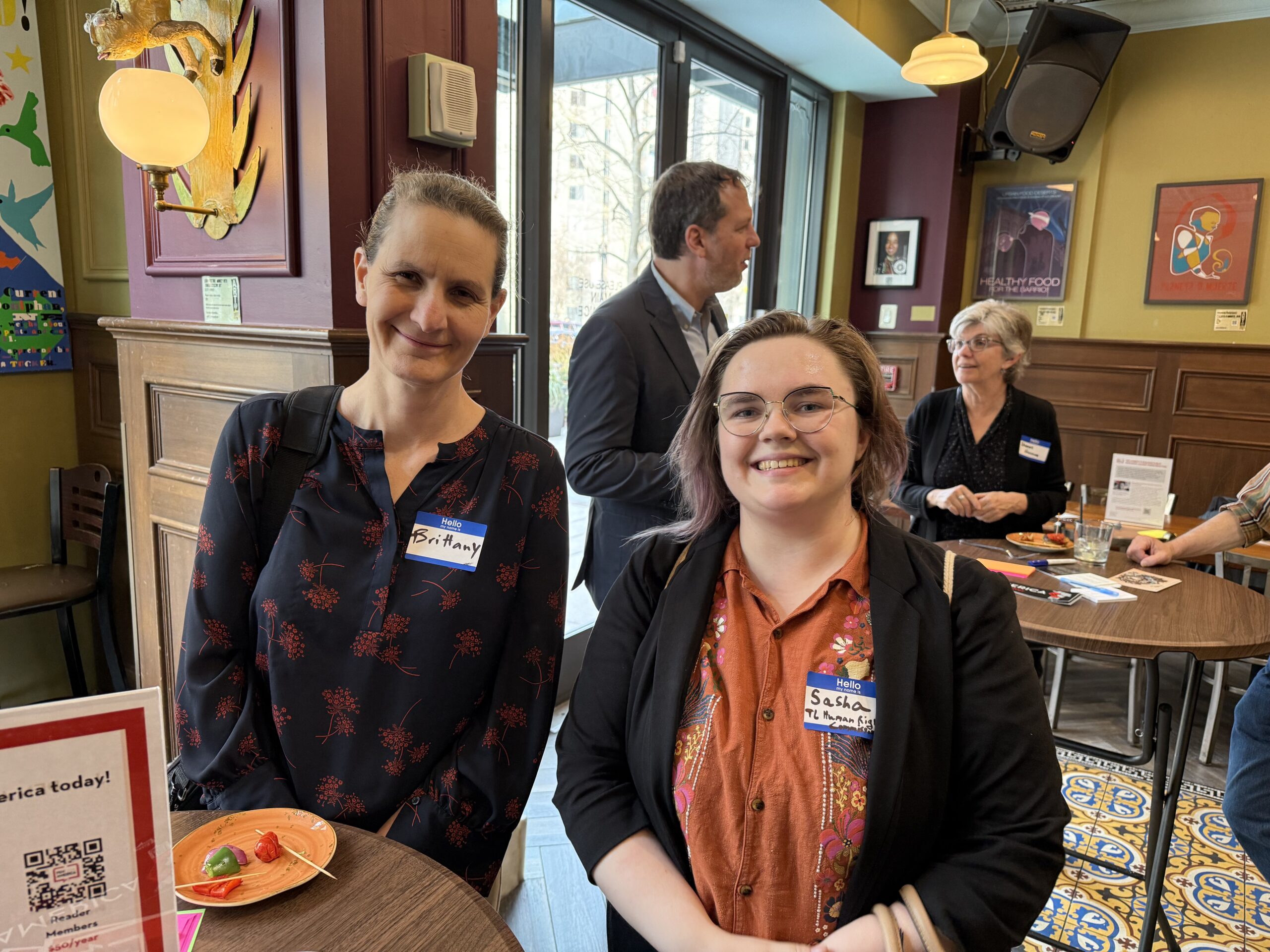 Two smiling people stand in a cafe holding plates. Theyre wearing name tags. Behind them are other people chatting around tables. The room has large windows and colorful decorations.