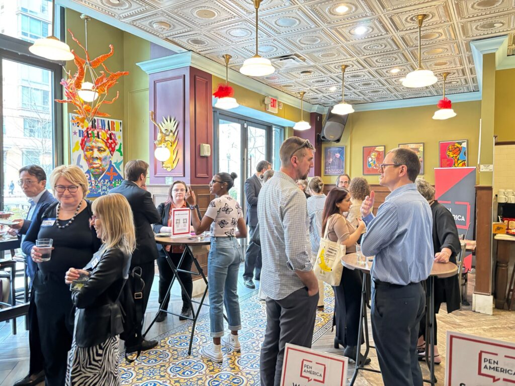 A diverse group of people socializing at an indoor gathering. There are colorful decorations and artwork on the walls. Attendees stand and chat in small groups, holding drinks and mingling around tables.