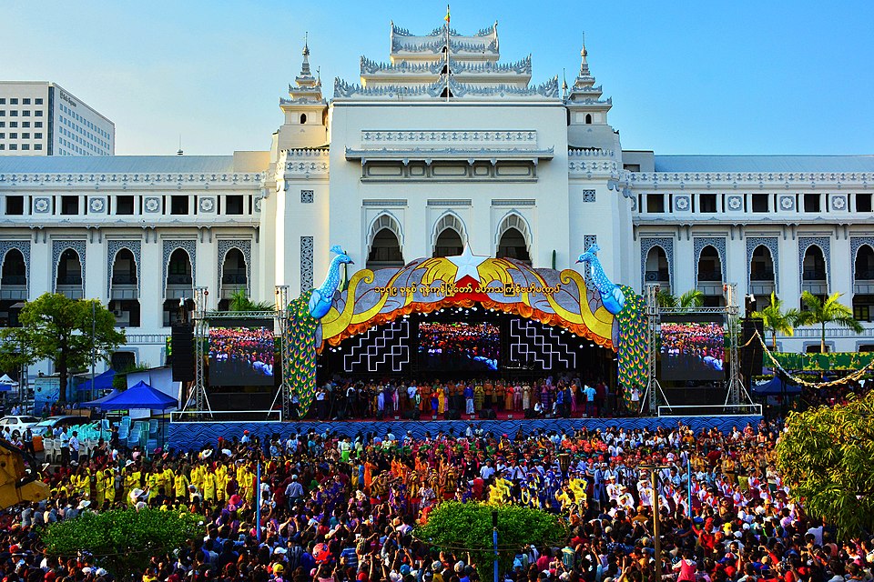 A large crowd gathers in front of an ornate white building with multiple turrets. A colorful stage is set up with performers and intricate decorations. The sky is clear, indicating a sunny day. Trees and banners add to the festive atmosphere.