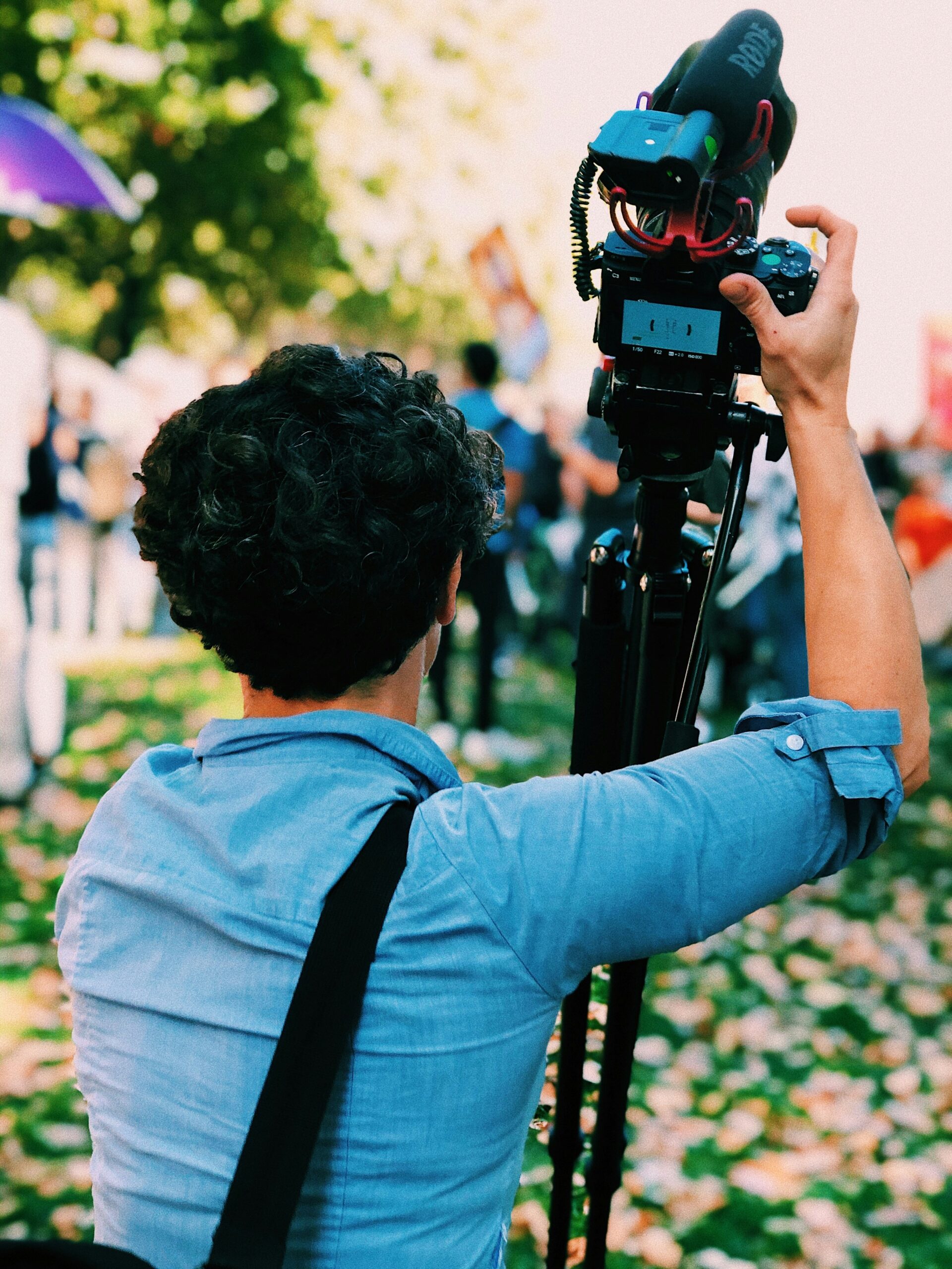 Person in a blue shirt with curly hair using a camera on a tripod outdoors. The ground is covered with fallen leaves, and there are blurred people and trees in the background, indicating an outdoor event or public gathering.