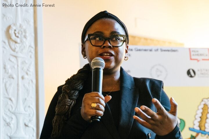A woman with glasses and braided hair speaks into a microphone. She is wearing a black suit. Behind her is a banner with text and logos. The setting is a well-lit room with decorative walls.