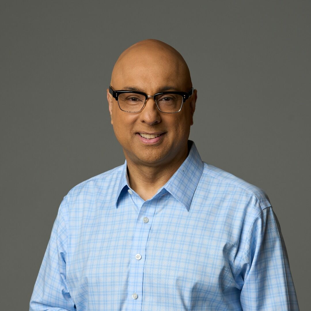 A person with a shaved head and glasses is smiling, wearing a light blue checkered shirt. The background is plain gray.