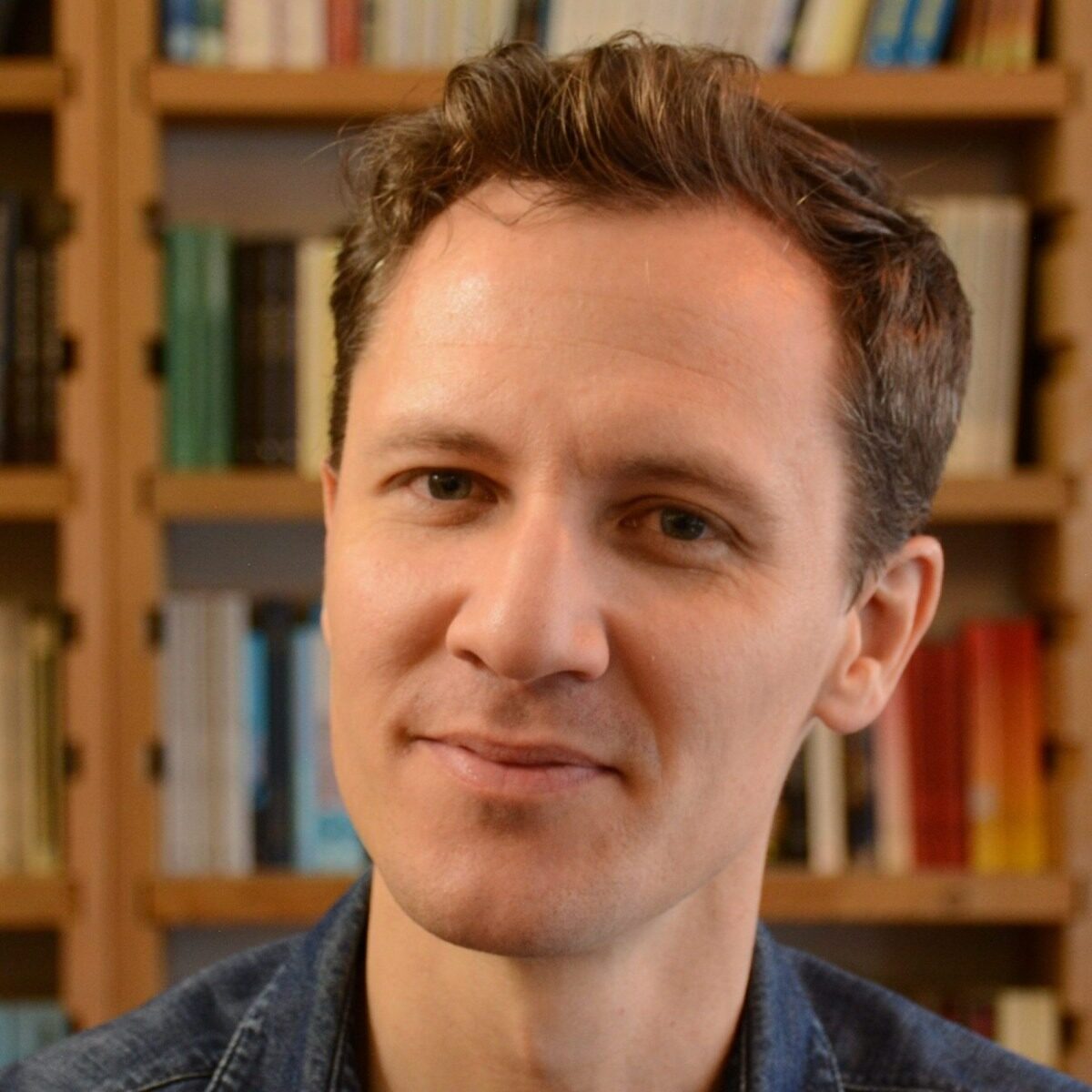 A person with short, brown hair is smiling slightly at the camera. They are wearing a blue shirt. Behind them is a blurred background with wooden shelves filled with assorted books.