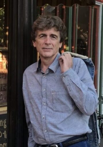 A man with curly hair wearing a blue checkered shirt and jeans leans against a black pillar. He has a jacket draped over his shoulder and stands outside near a storefront with glass windows.