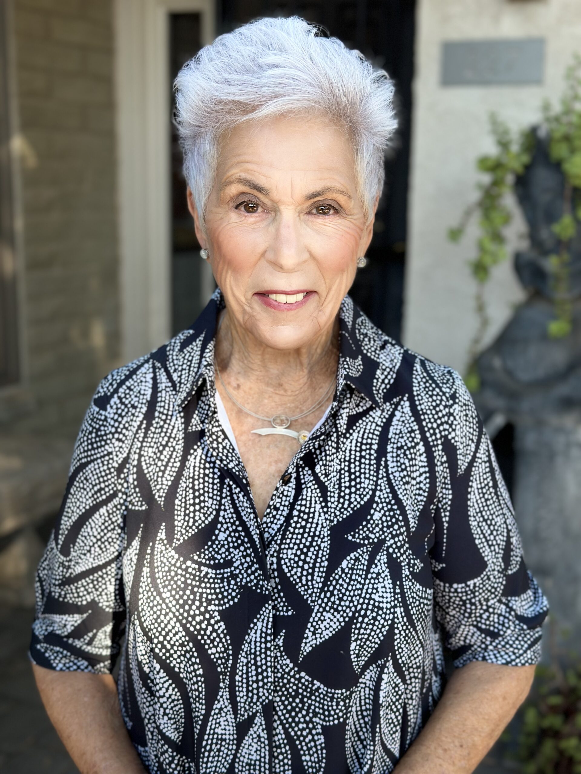 An older woman with short white hair smiles while standing outdoors. She is wearing a patterned black and white blouse. Theres a building wall in the background with some greenery.