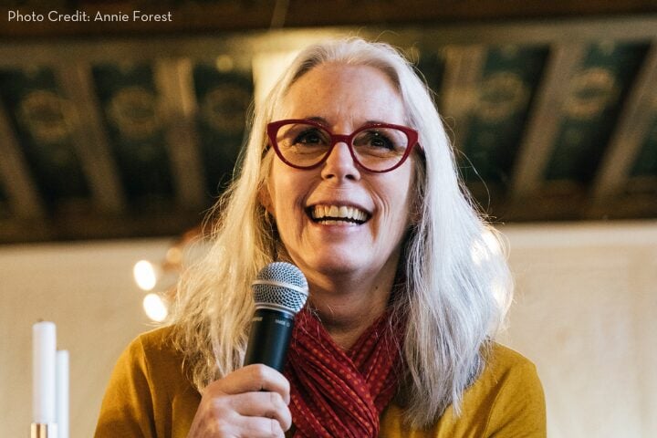A smiling woman with long gray hair and red glasses holds a microphone. She wears a red scarf and a mustard yellow top. The background is softly blurred with decorative elements.
