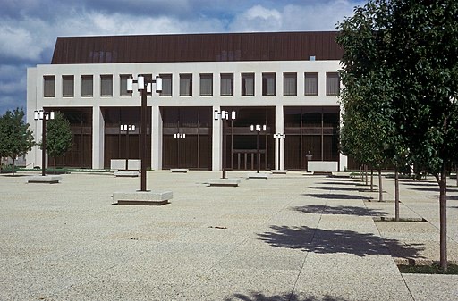 A large, modernist building with a flat roof and white facade, featuring a series of tall, narrow windows. The foreground displays a plaza with patterned paving, trees, and unique lamp posts under a partly cloudy sky.