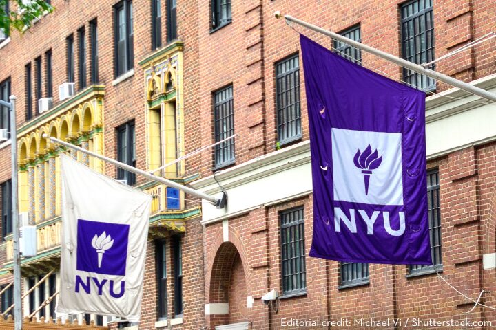 Two flags displaying the NYU logo hang from a brick building. One flag is purple with a white torch and NYU text, and the other is white with a purple torch and NYU text. The building has decorative architectural elements.