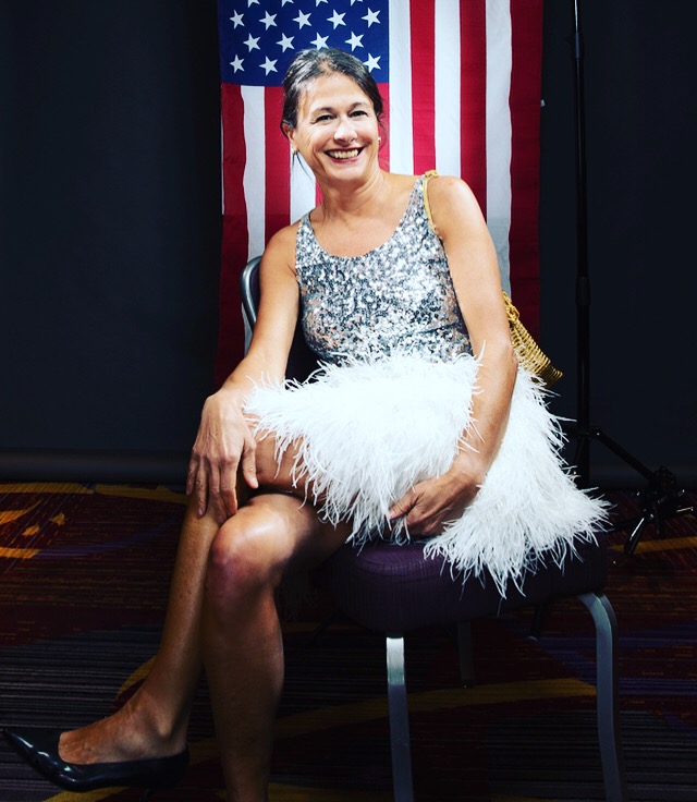 A woman in a silver sequin top and white feathered skirt sits smiling on a chair in front of a hanging American flag. The background is dark and the setting appears to be indoors.
