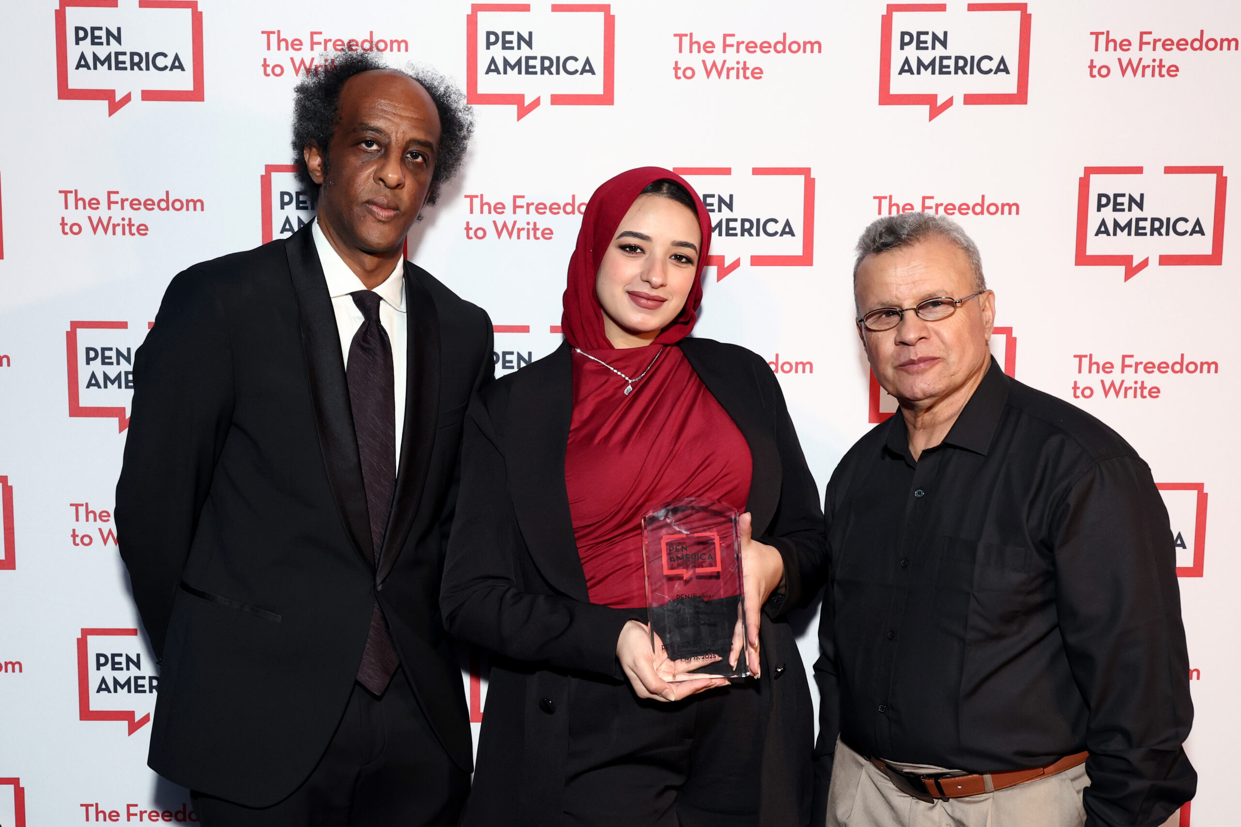 Three people stand together at a PEN America event. The woman in the center, wearing a red hijab and holding an award, is flanked by two men. The background features the PEN America logo and “The Freedom to Write” slogan.