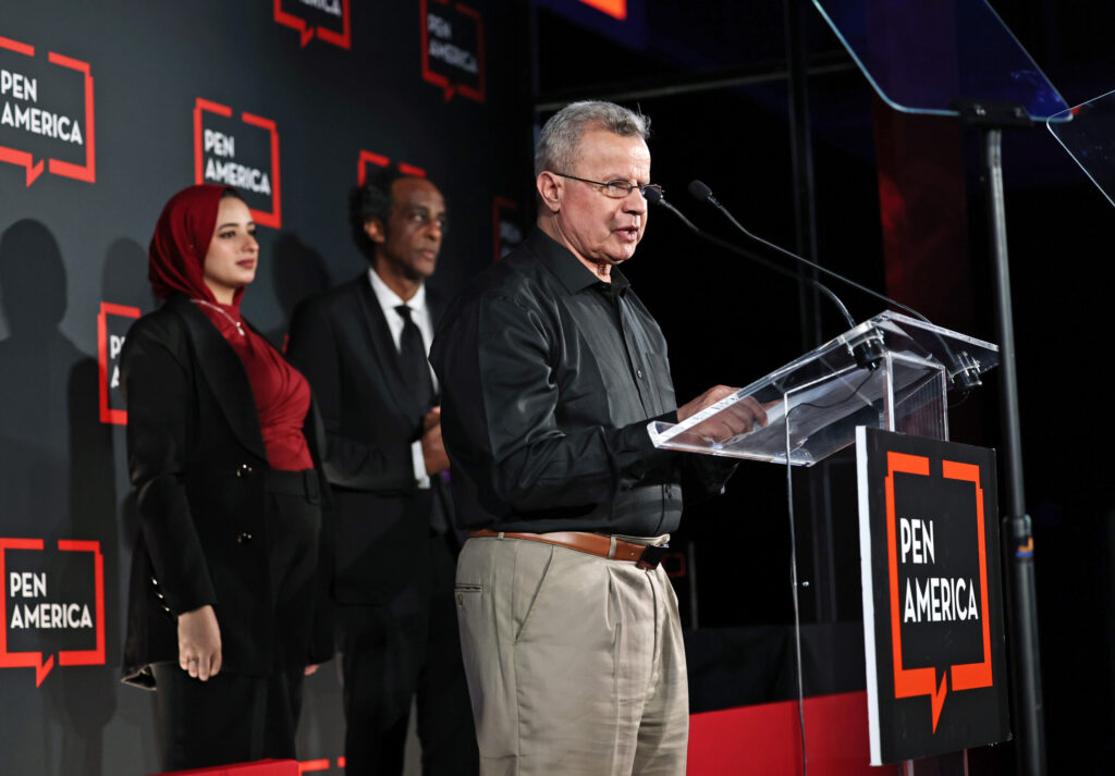 A man speaks at a clear podium with a PEN America sign, while two people stand behind him on stage. The background displays multiple PEN America logos on black panels.