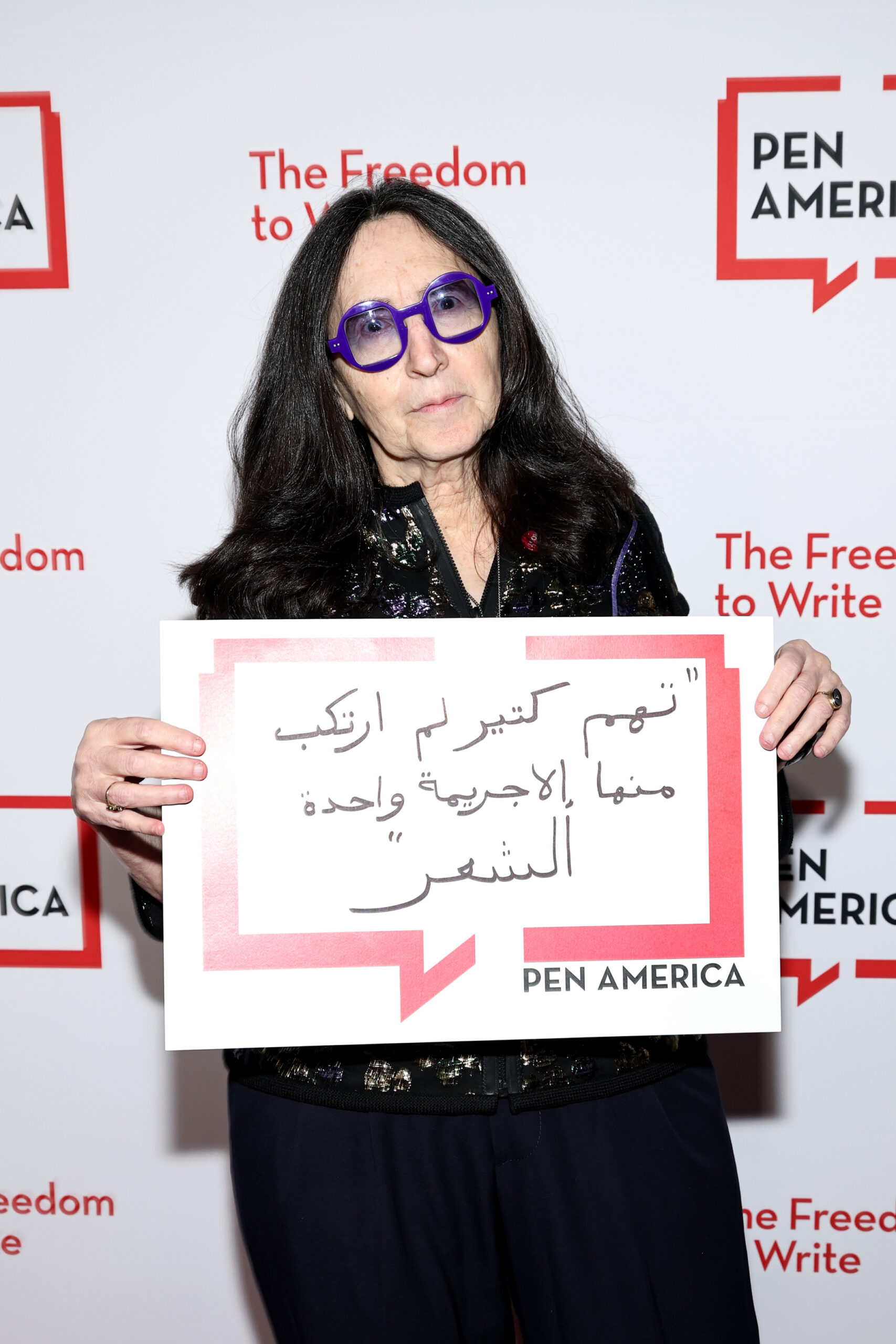 A woman with long dark hair and sunglasses holds a sign with Arabic text in front of a PEN America backdrop. The backdrop reads The Freedom to Write and PEN America in red and black.