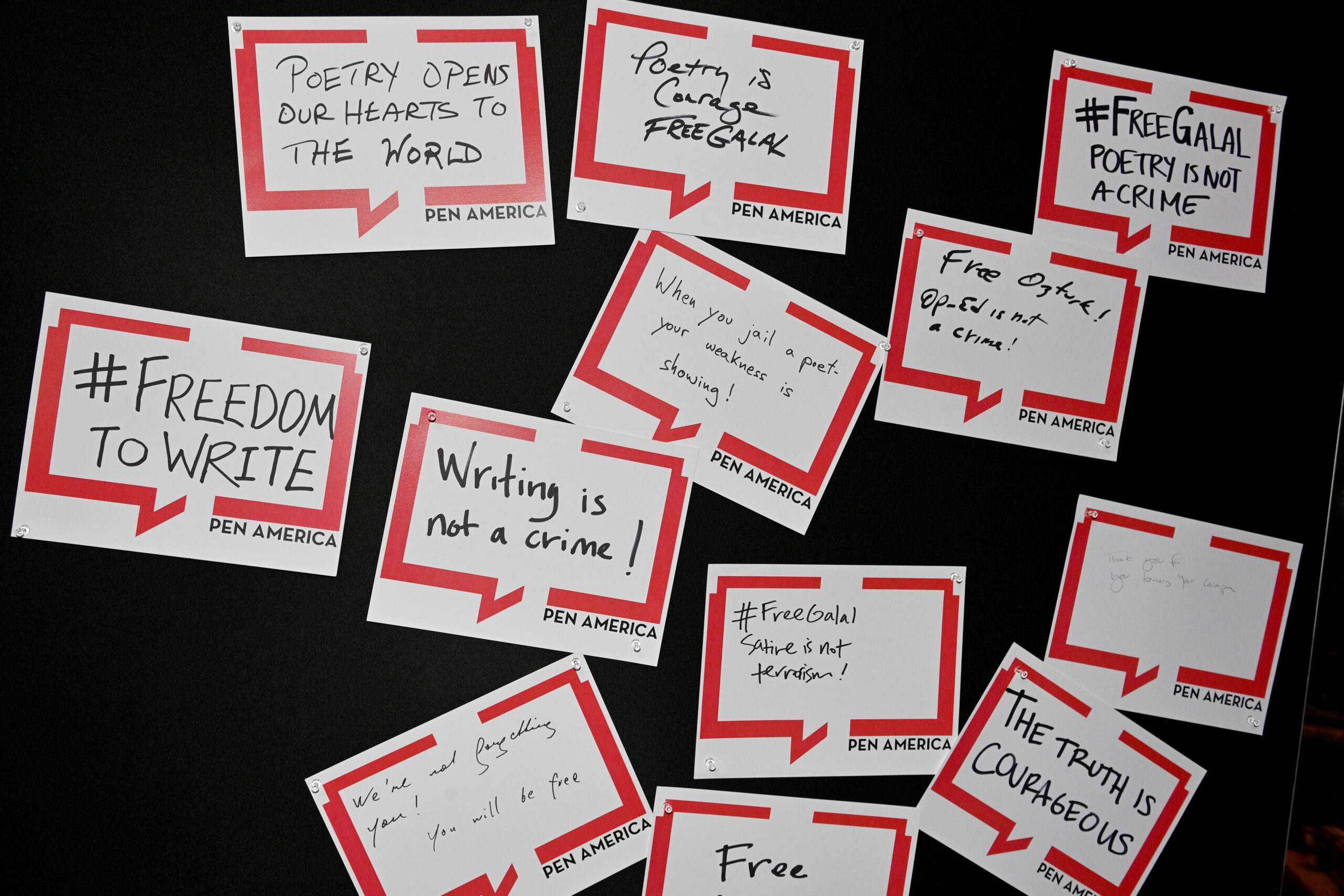 A collection of handwritten signs with messages like “#FREEDOMTOWRITE,” “Writing is not a crime!,” “Poetry opens our hearts to the world,” and “The truth is courageous,” displayed on a black background.