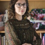 A woman with wavy brown hair and glasses stands with arms crossed, wearing a patterned blouse. She is in front of a bookshelf filled with books and a floral painting.