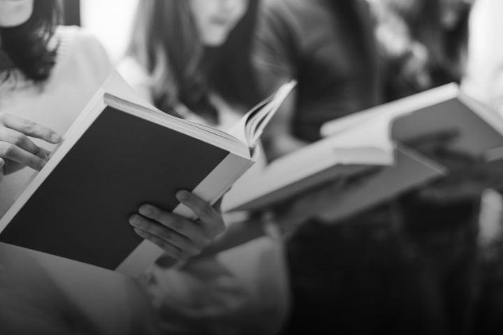 A black and white photo showing a group of people holding and reading books, with their faces partially out of focus or cropped out of the frame.