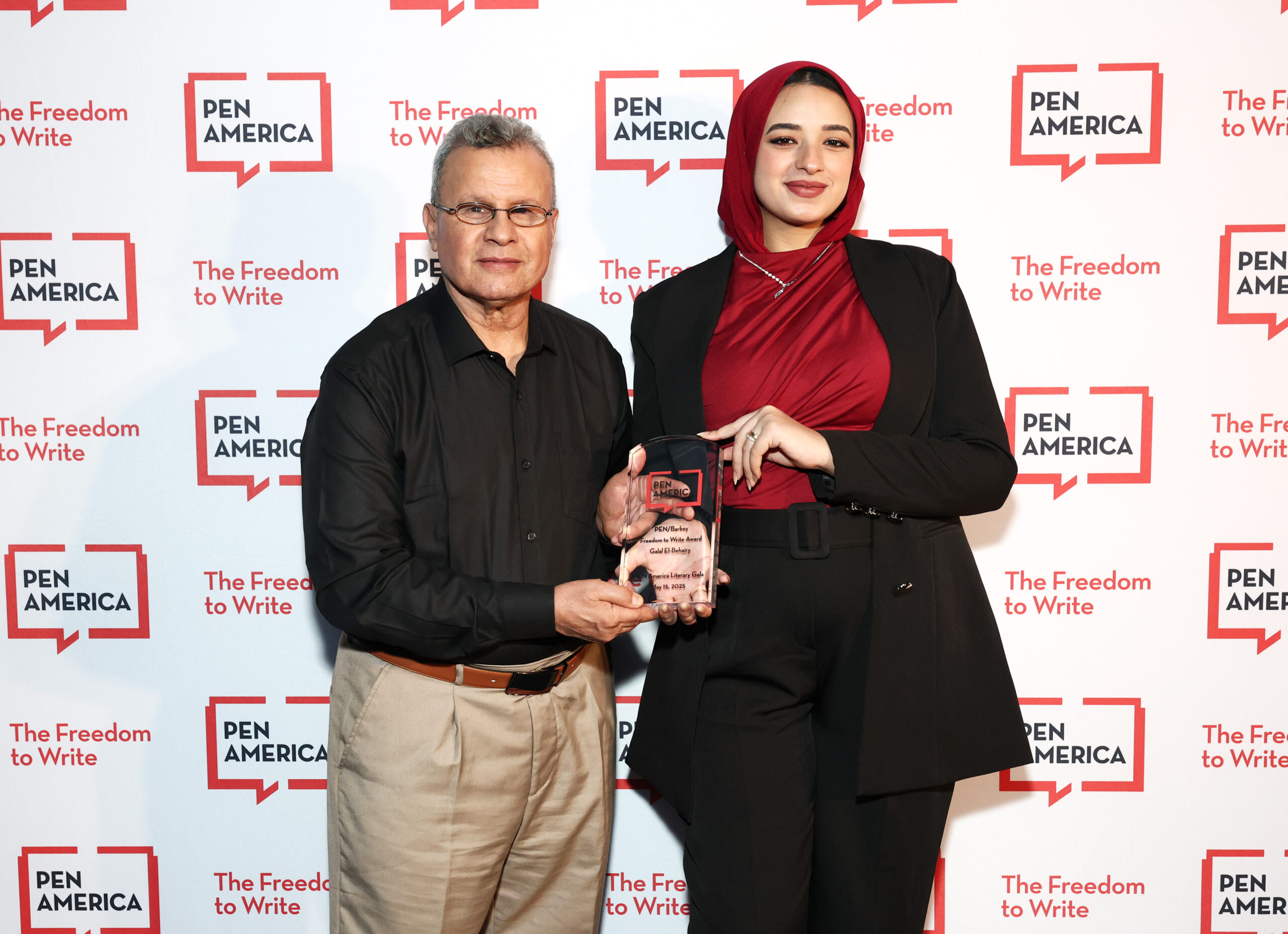 Two people stand together holding an award in front of a PEN America backdrop. One person wears a black shirt and khaki pants, the other wears a red hijab, black blazer, and red top. Both are smiling at the camera.