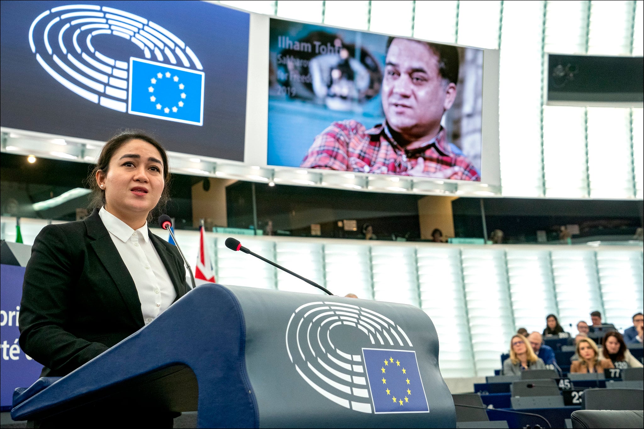 A woman speaks at a podium with the European Union emblem in a large assembly hall. Behind her, a screen displays a man’s portrait and the name Ilham Tohti. Seated audience members are visible in the background.