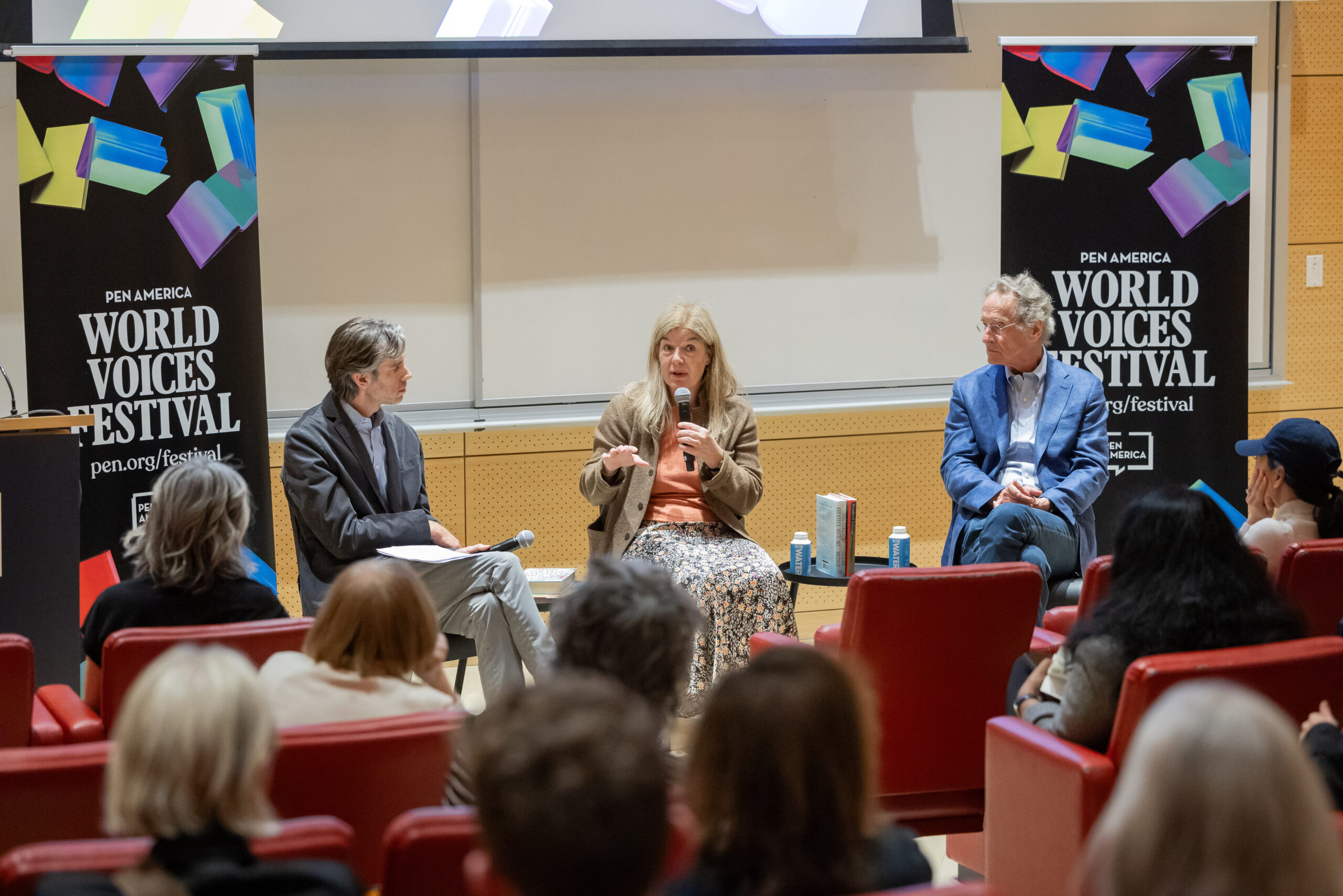 Three speakers sit at the front of a room, engaging in a discussion at the PEN America World Voices Festival. The audience listens attentively, and colorful festival banners are displayed on both sides of the stage.