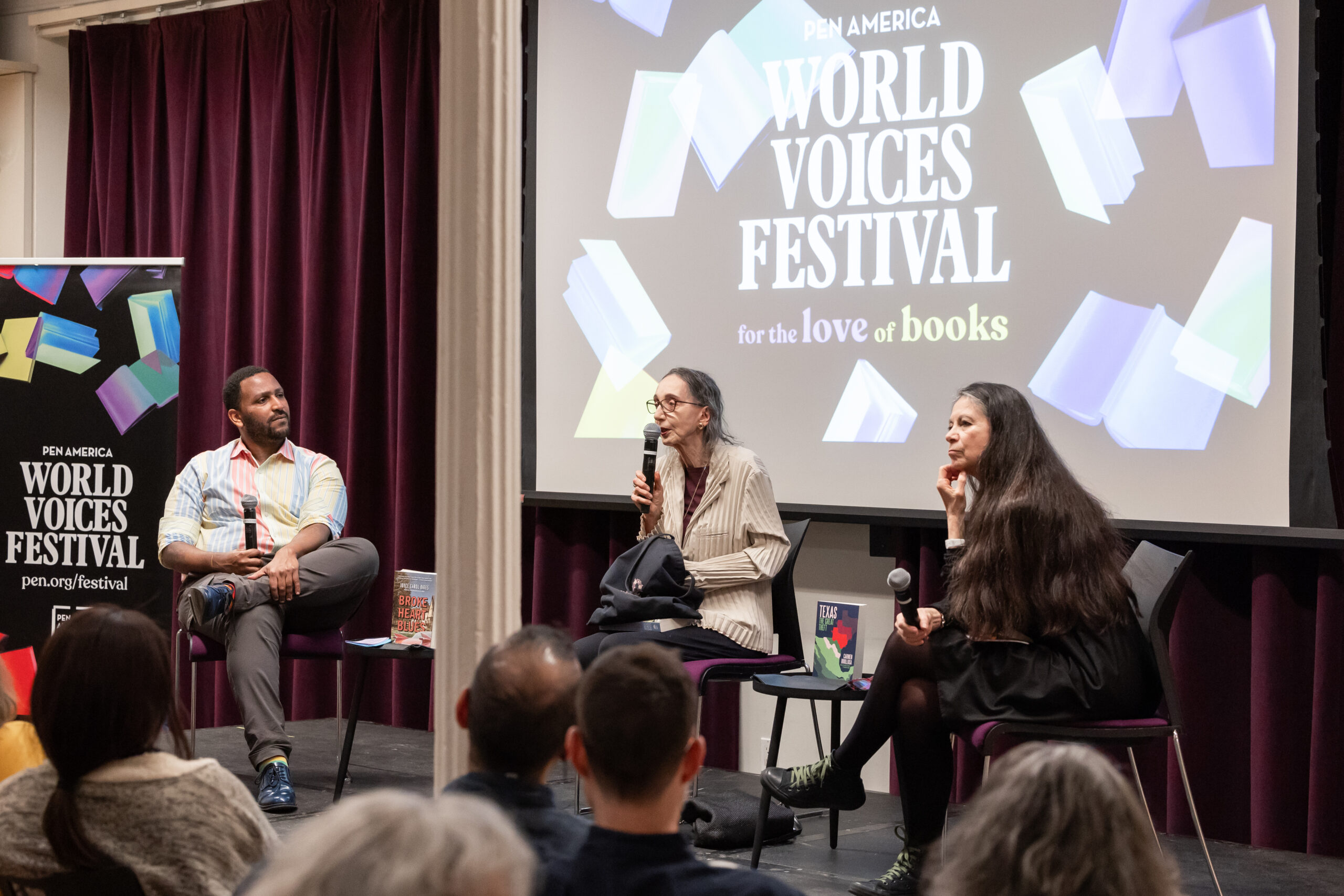 Three speakers sit on stage during a panel discussion at the PEN America World Voices Festival, with an audience watching. A large screen behind them displays the event name, World Voices Festival: for the love of books.