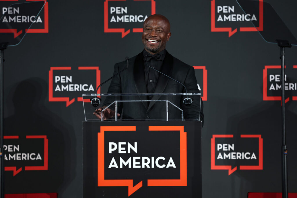 A man in a black suit smiles while speaking at a podium with a PEN America sign. The background features multiple PEN America logos on a dark backdrop.
