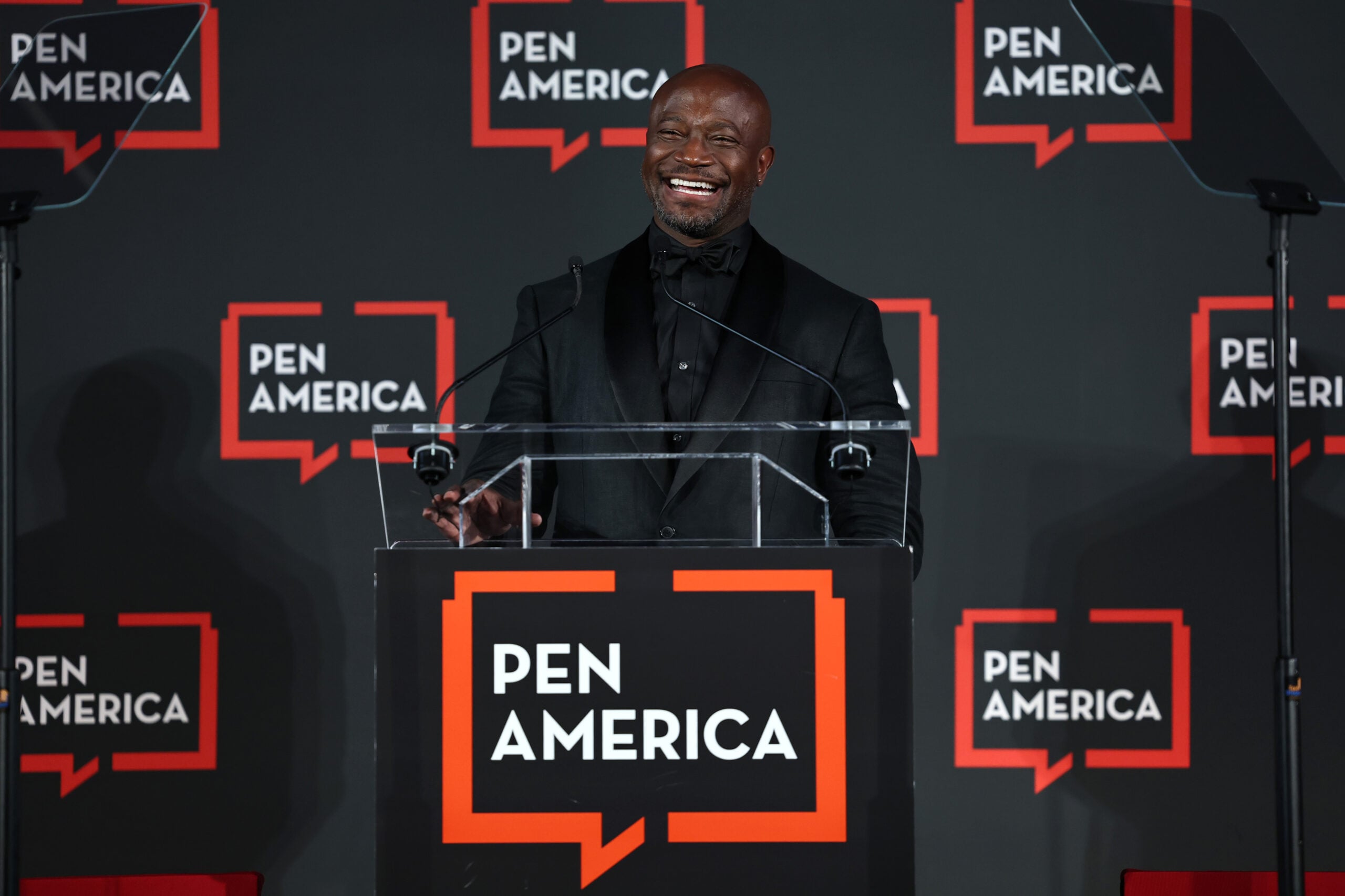 A man in a black suit smiles while speaking at a podium with a PEN America sign. The background features multiple PEN America logos on a dark backdrop.
