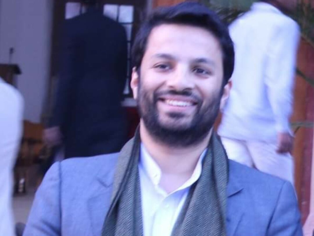 A man with a beard smiles while standing outdoors, wearing a gray suit, white shirt, and green scarf. The background shows people in white attire and blurred architectural details.