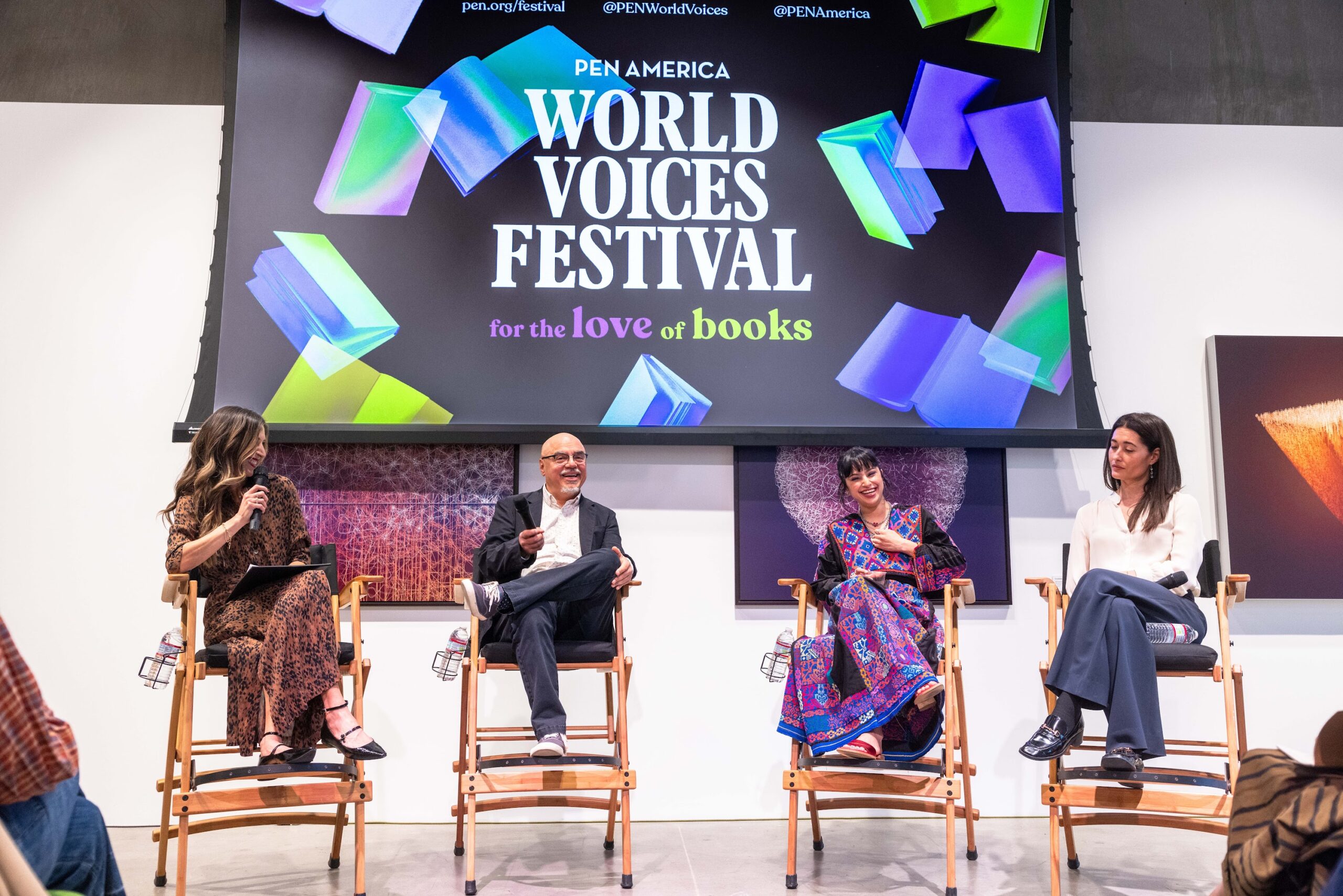 Four panelists sit on stage in front of a colorful PEN America World Voices Festival for the love of books sign, engaging in a discussion, with microphones and notes in hand.