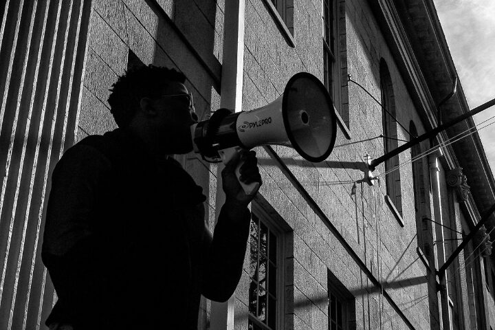 A person holds a megaphone, speaking or shouting, standing next to a building with tall columns. The image is in black and white, with strong shadows and sunlight on the wall.
