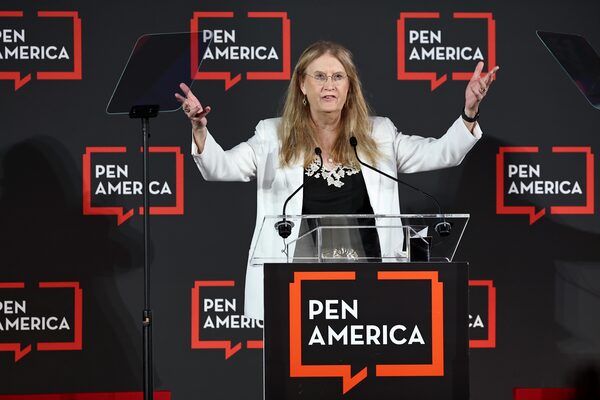A person with long hair wearing glasses and a white blazer speaks at a clear podium with PEN America signs in the background, gesturing with raised arms during an event.