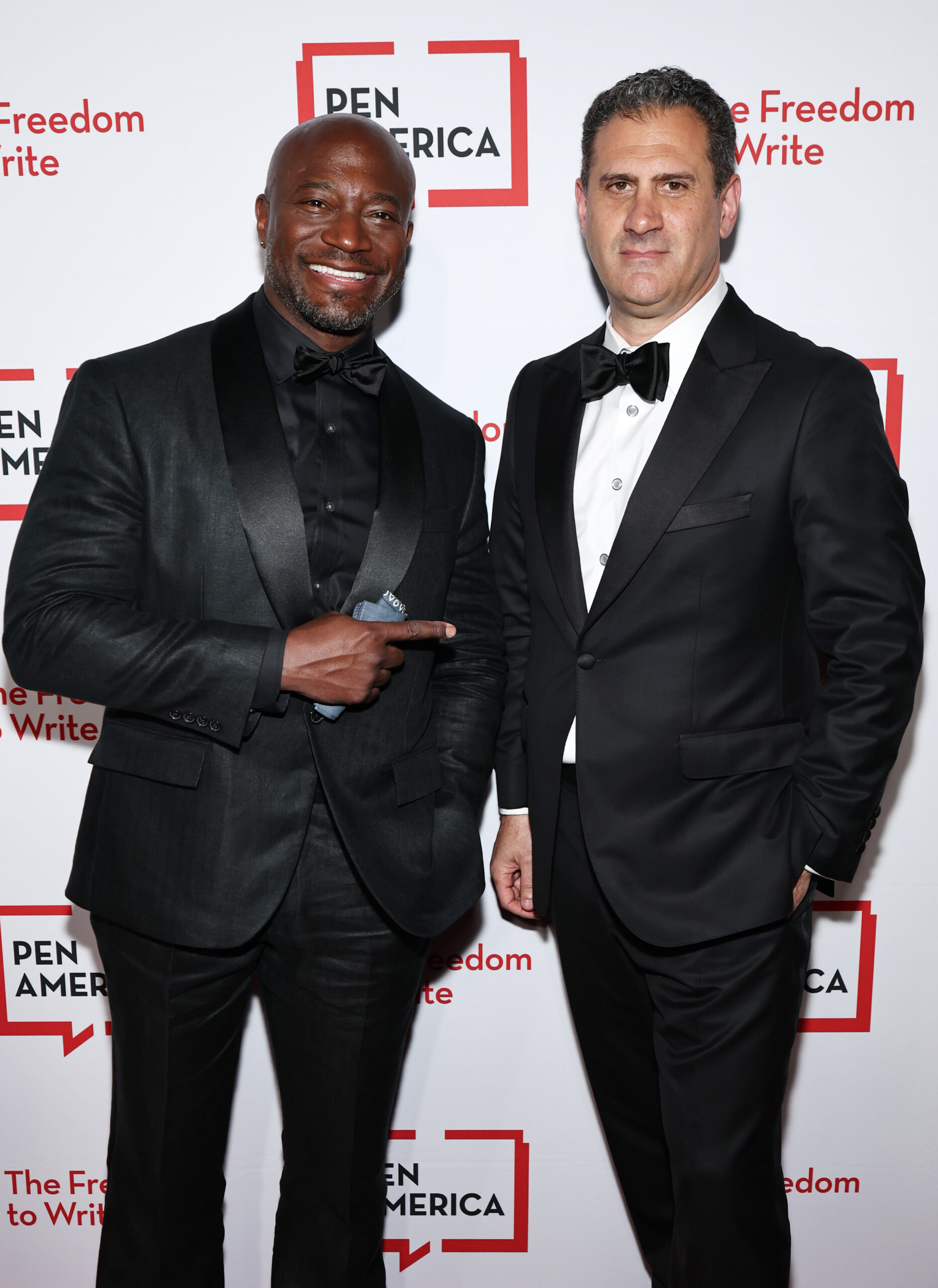 Two men in black tuxedos pose and smile at a PEN America event, standing in front of a white backdrop with red and black PEN America logos and the words “The Freedom to Write.” One man points toward the other.