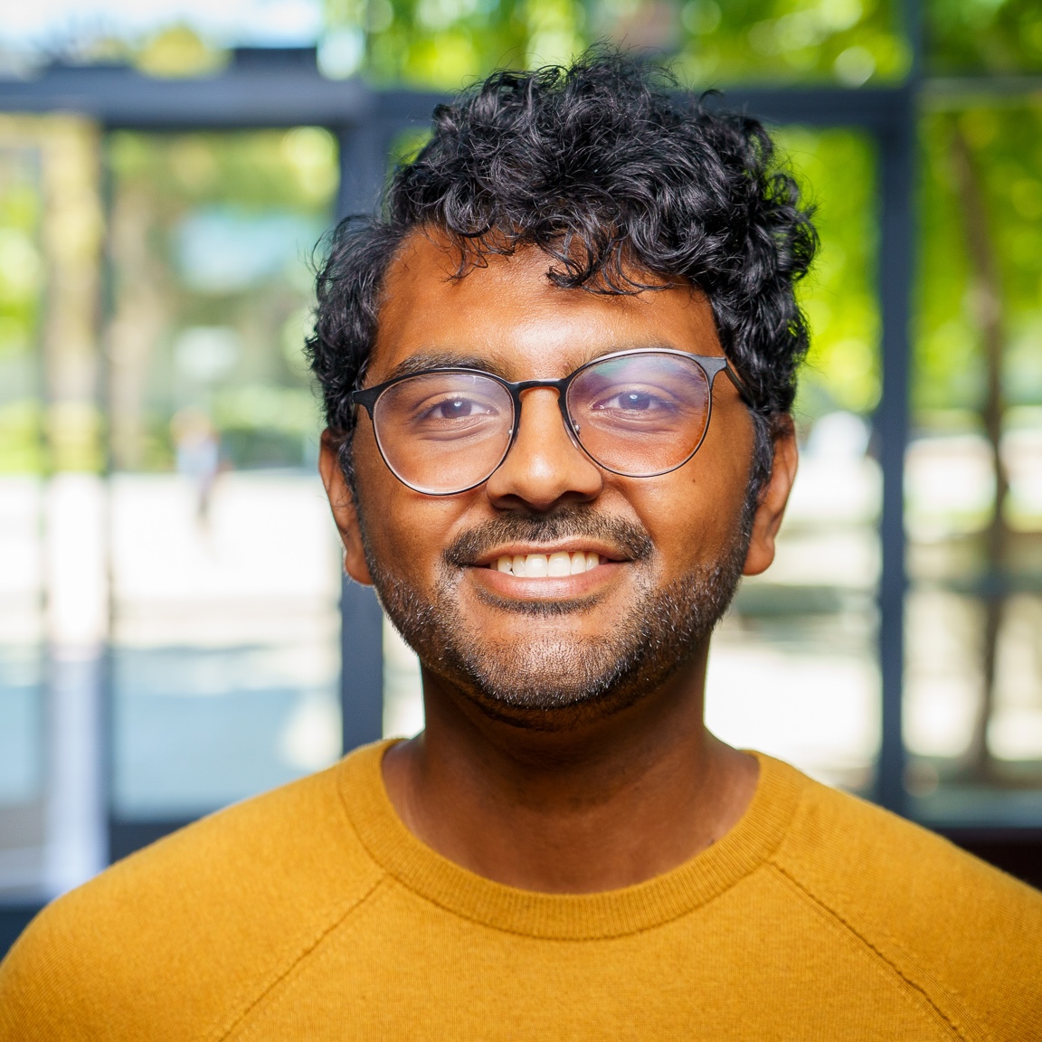 A person with short curly hair, glasses, and a beard smiles while wearing a mustard yellow sweater. They stand indoors with sunlight and greenery visible through large windows behind them.