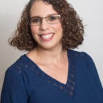 A woman with curly brown hair and glasses smiles at the camera. She is wearing a navy blue blouse with lace trim and stands against a plain light background.