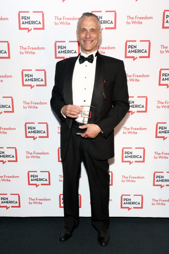 A smiling man in a black tuxedo holds an award while standing in front of a PEN America step-and-repeat backdrop with the slogan “The Freedom to Write.”.